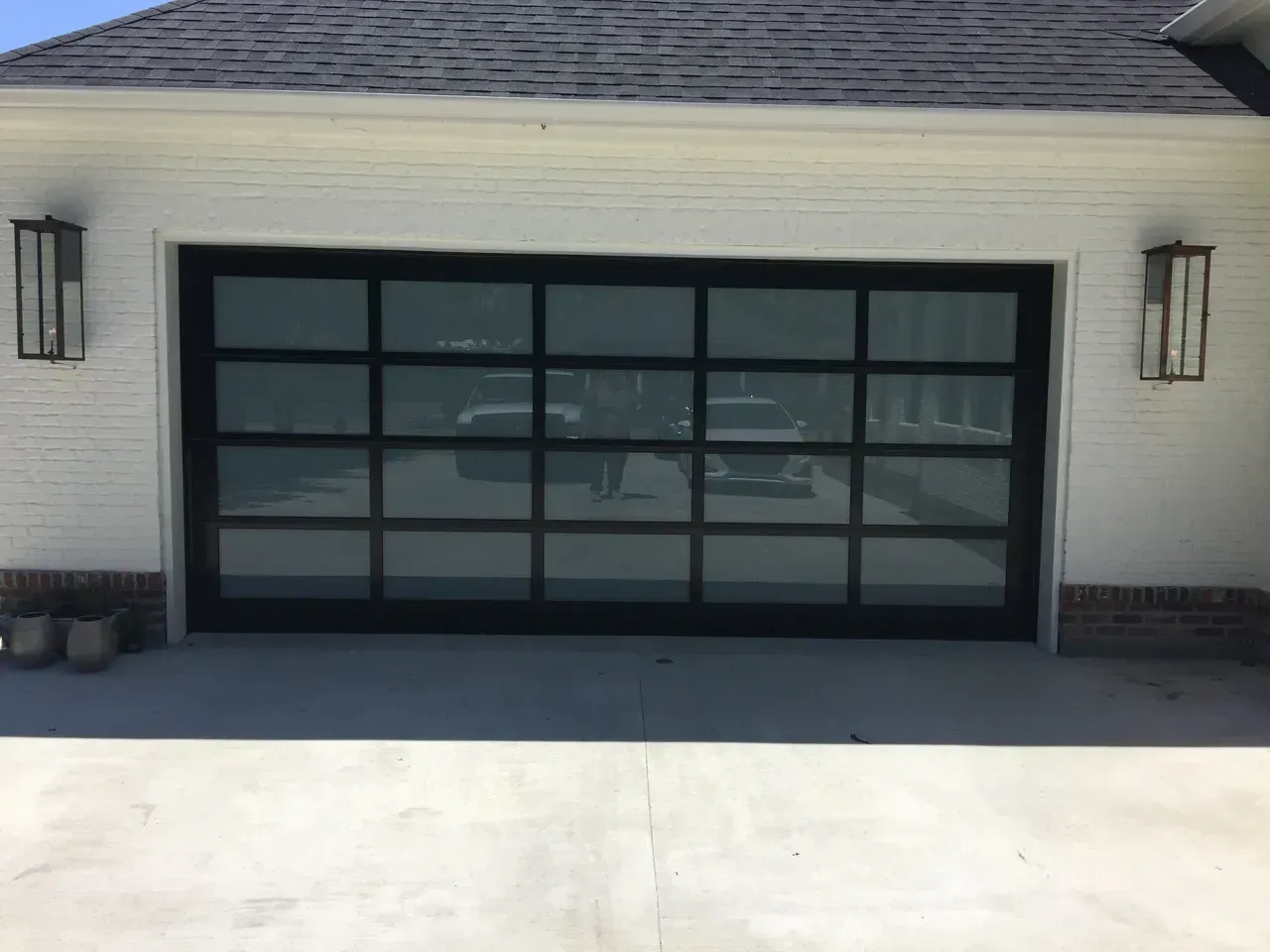 Black-framed glass garage door reflects vehicles and other objects.  White brick building with two sconces.