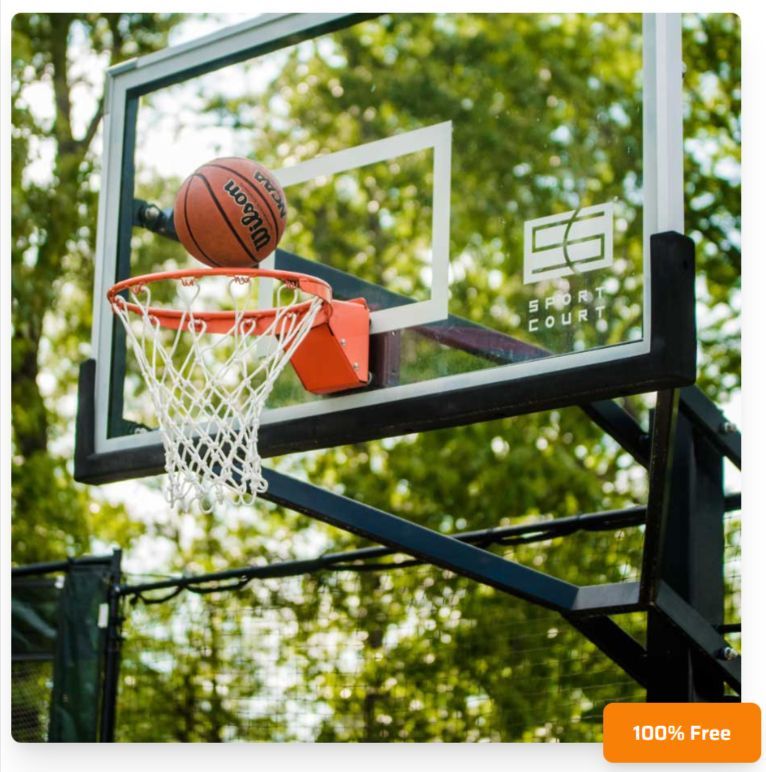 A Wilson basketball sits on the rim of an outdoor basketball hoop with a clear backboard and white net against green trees.