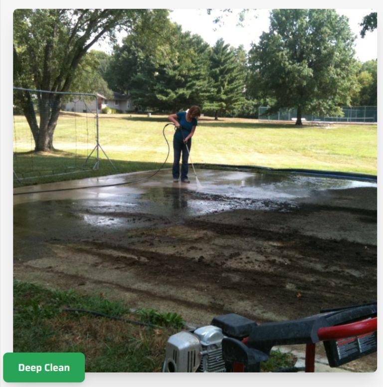 A person uses a pressure washer to clean a dark, dirty patch of concrete in a park setting.