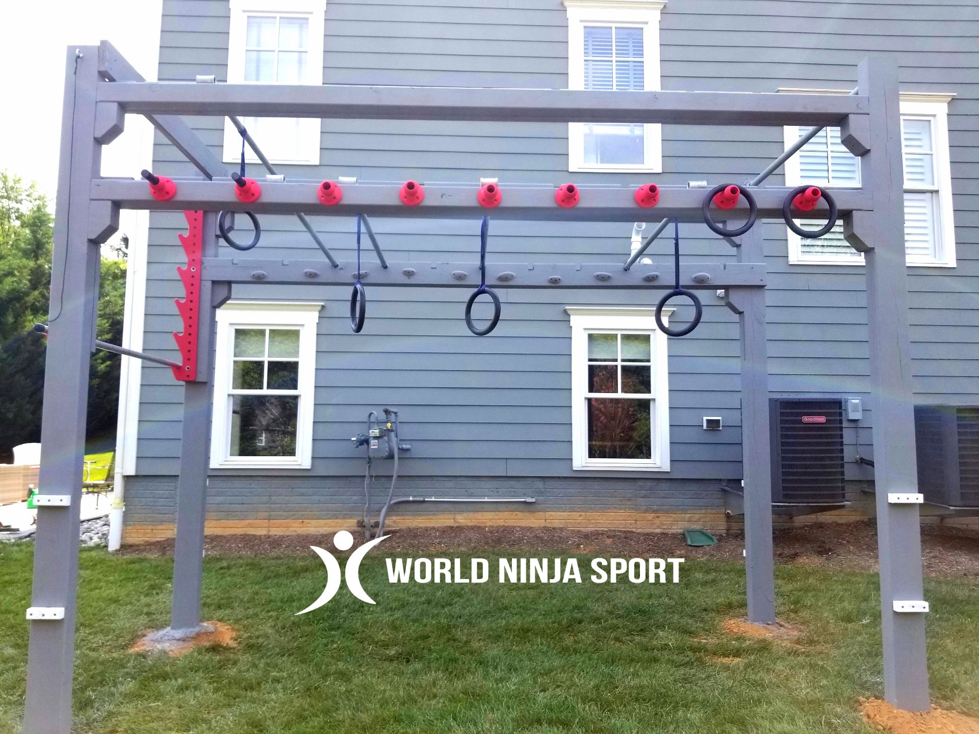 A grey outdoor wooden ninja obstacle course with hanging rings, mounted in front of a house on a lawn.