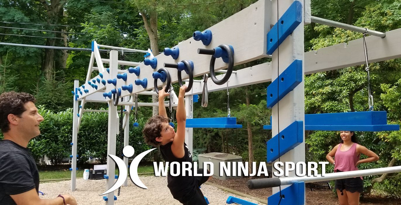 A child hangs from rings on an outdoor obstacle course, with a man supervising and another person standing nearby.