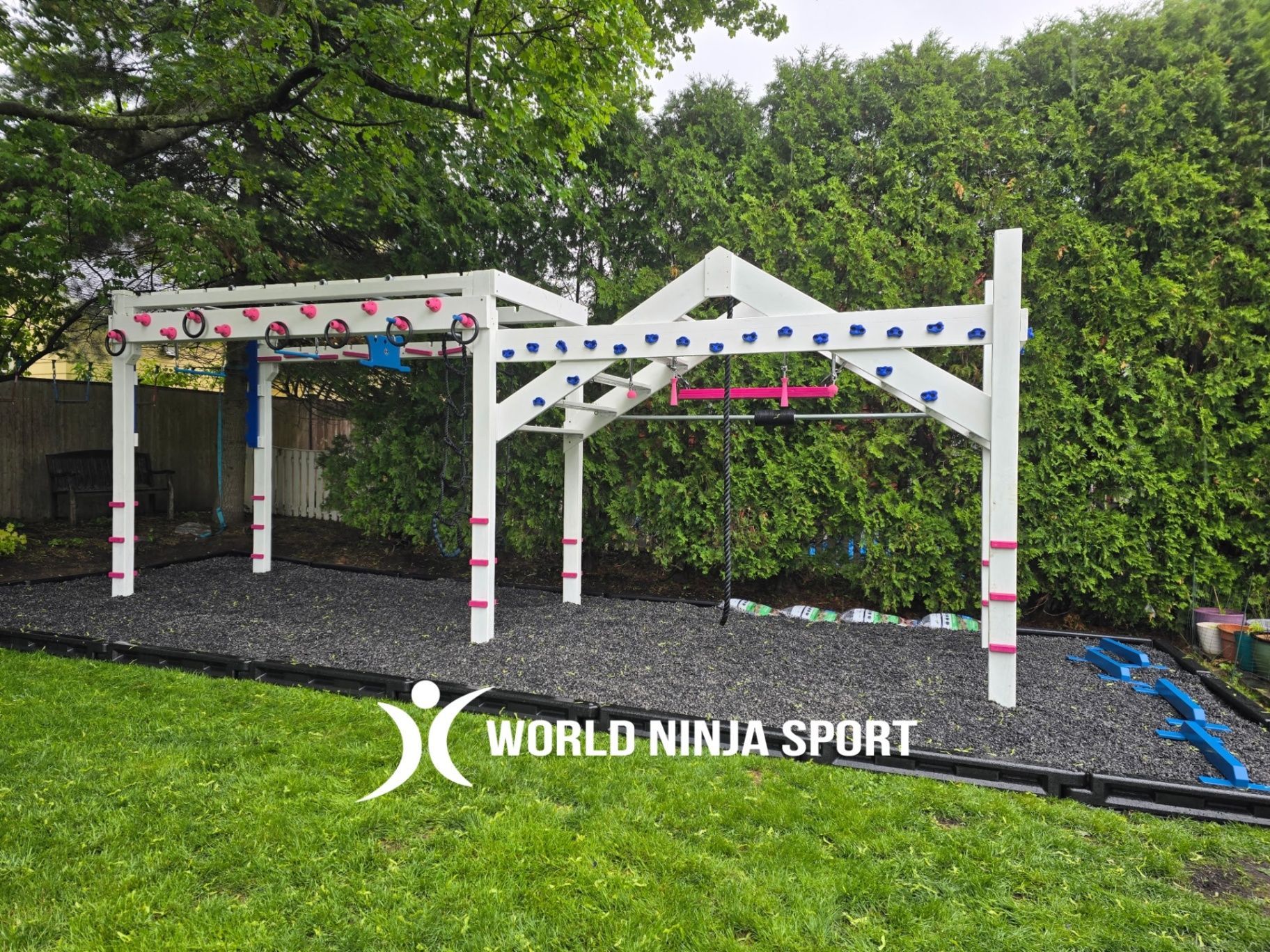 Outdoor wooden ninja obstacle course in a backyard, featuring bars and holds on a bed of black mulch.