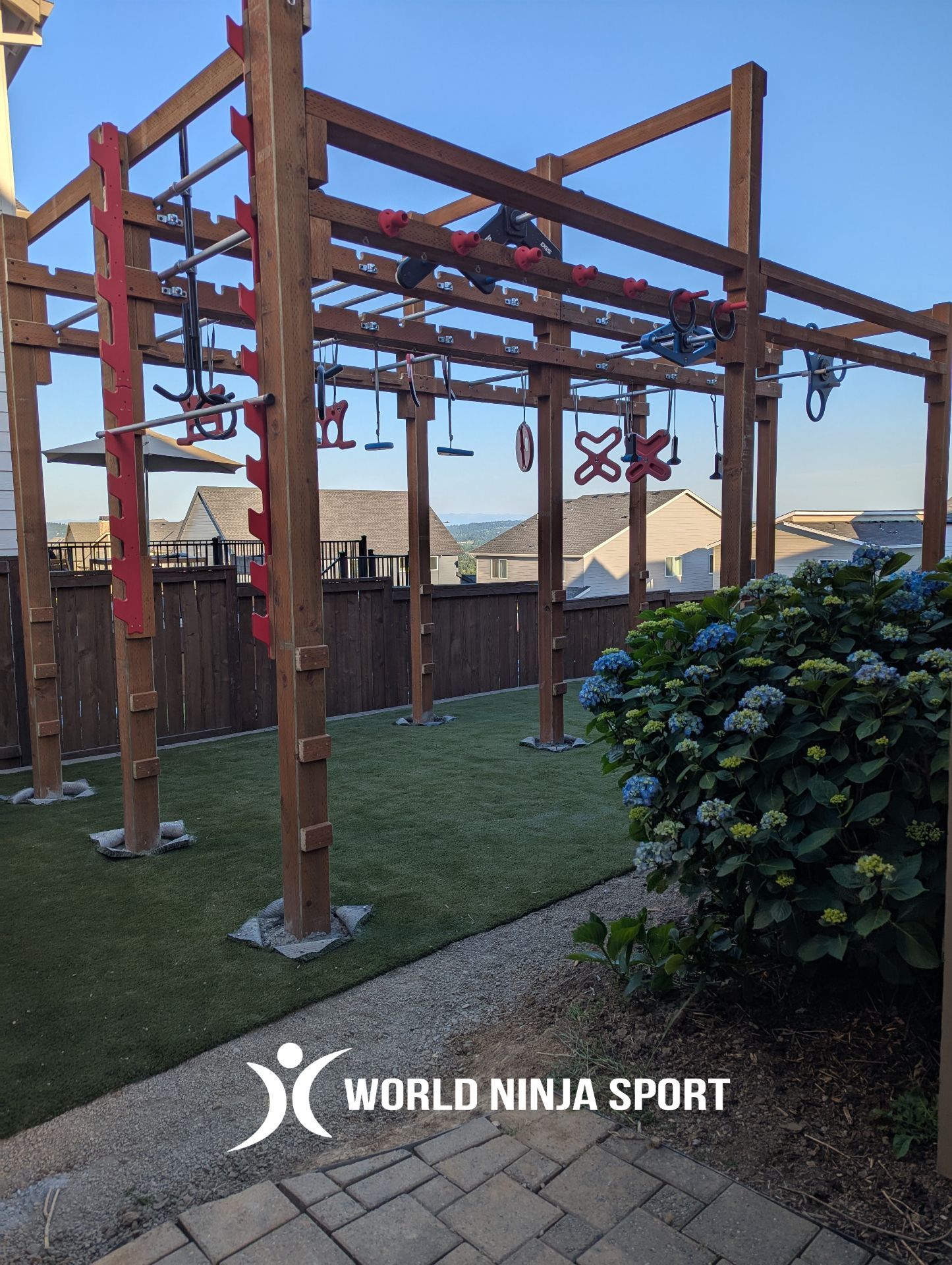 Outdoor wooden ninja warrior obstacle course structure with various hanging training grips, set on green grass.