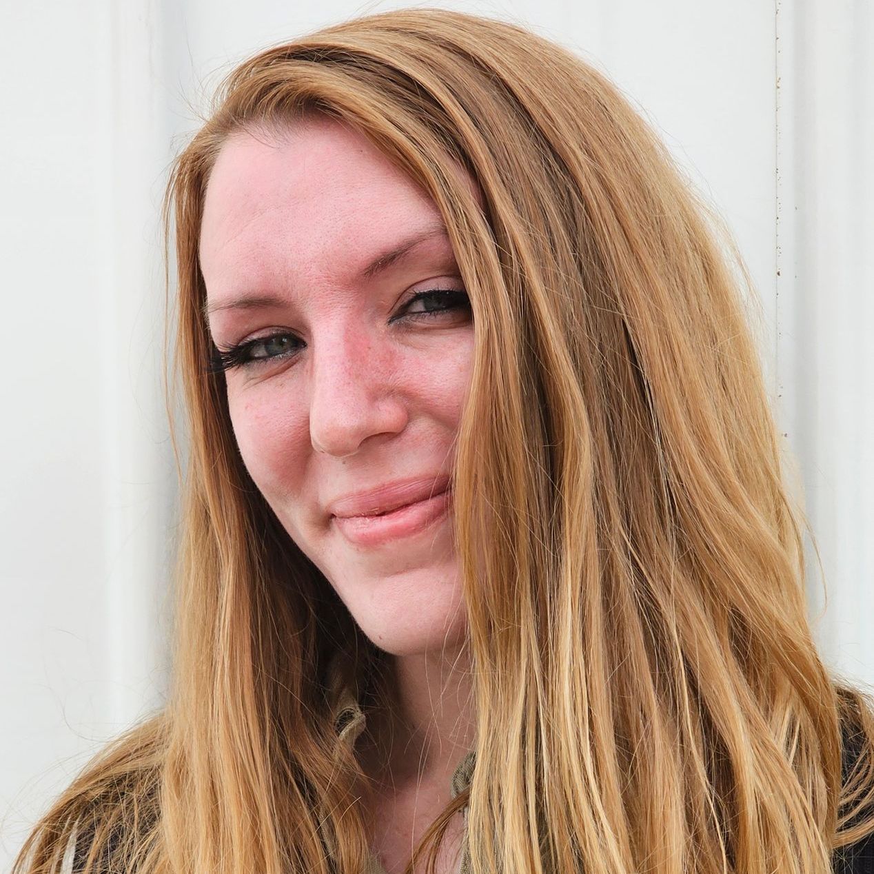 A woman with long blonde hair is smiling in front of a white wall.