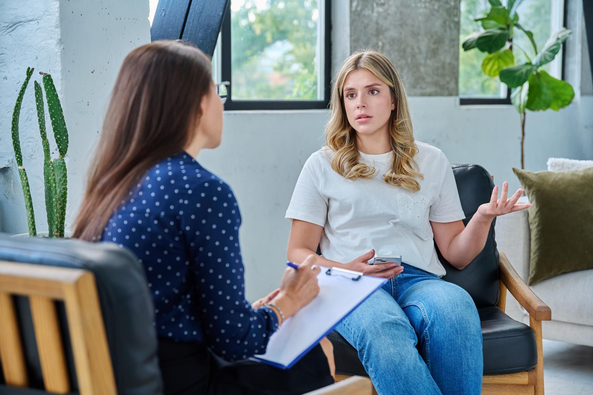 Two women in a counseling session, seated with clipboards in a bright room.