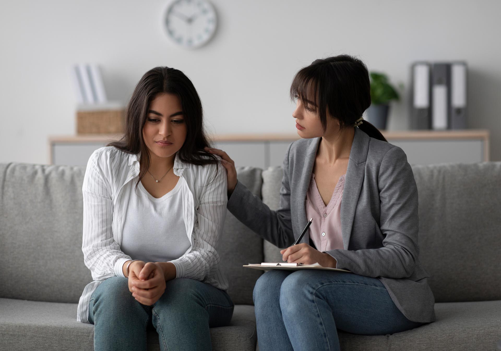 Two women sit on a gray couch in a counseling session, one offering support with a notebook in hand.