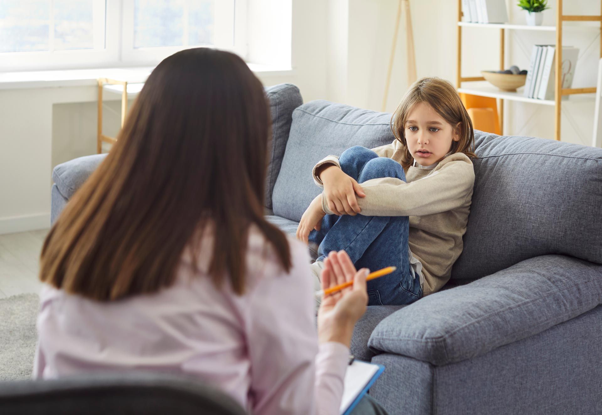 Two people talking on a gray sofa in a bright living room, one holding a phone