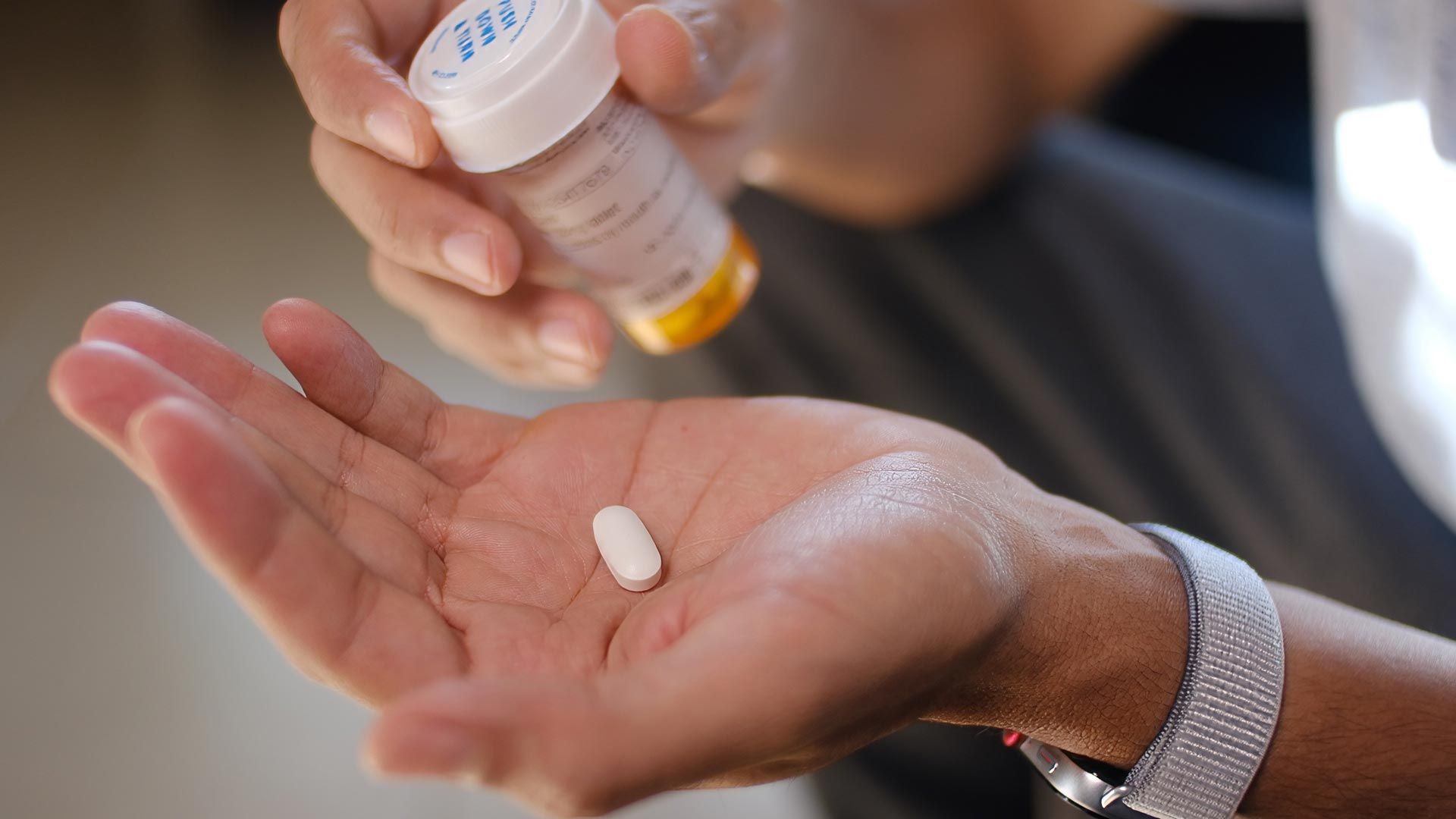 A hand holds a white tablet while another hand pours pills from a bottle above it.