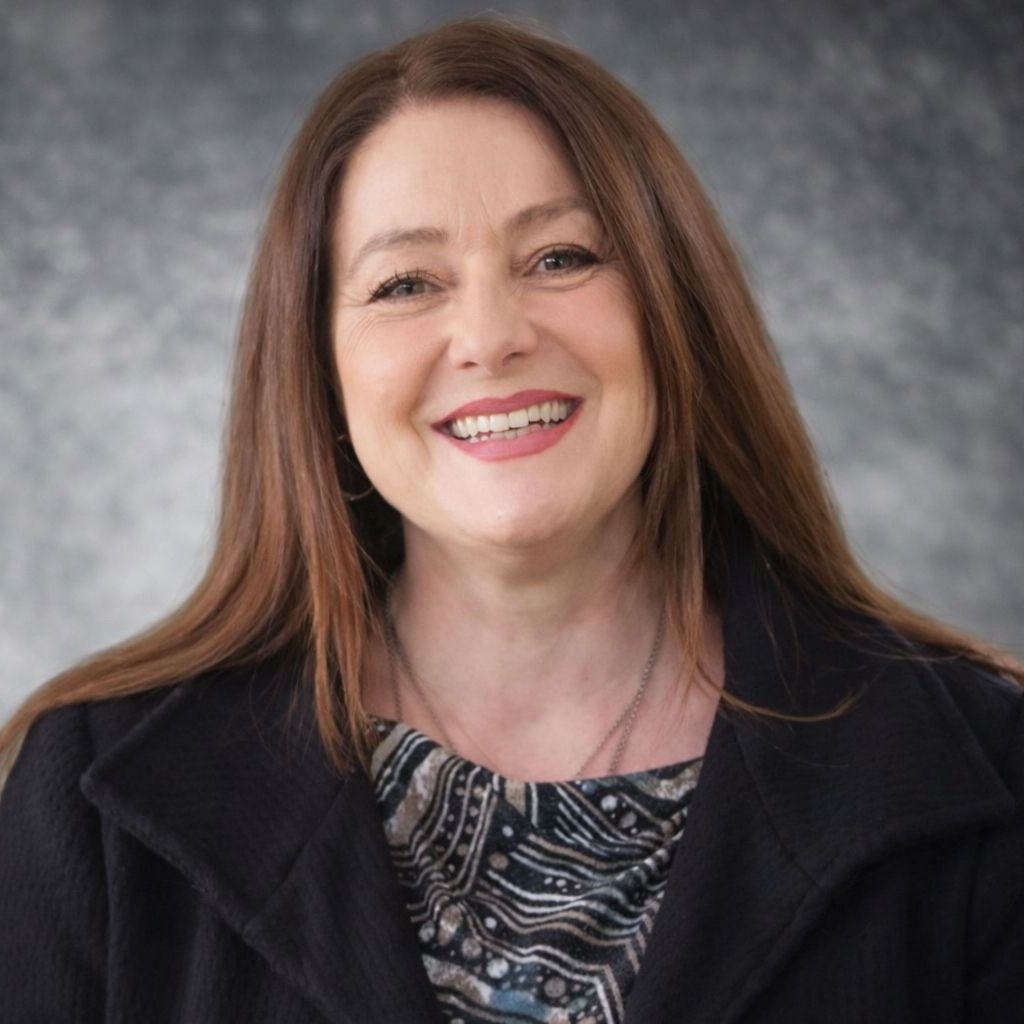 Woman with long brown hair smiles, wearing a black coat and patterned top, outdoors with greenery.