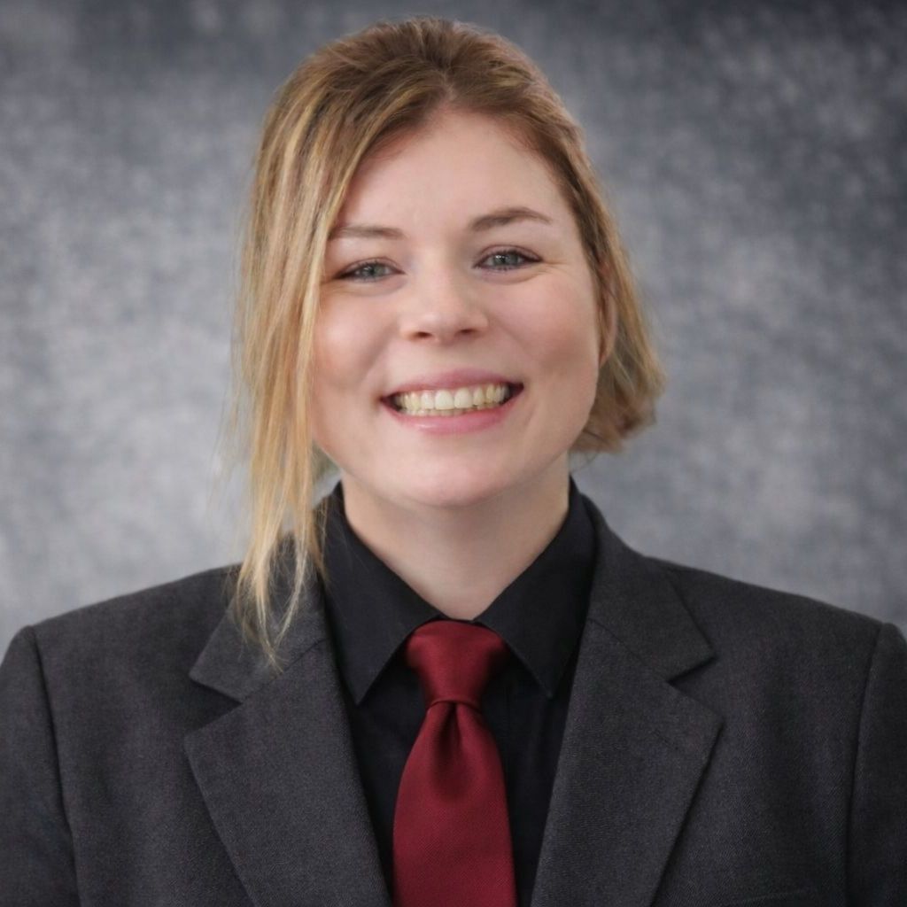 Woman in black suit smiles, standing outdoors near greenery.