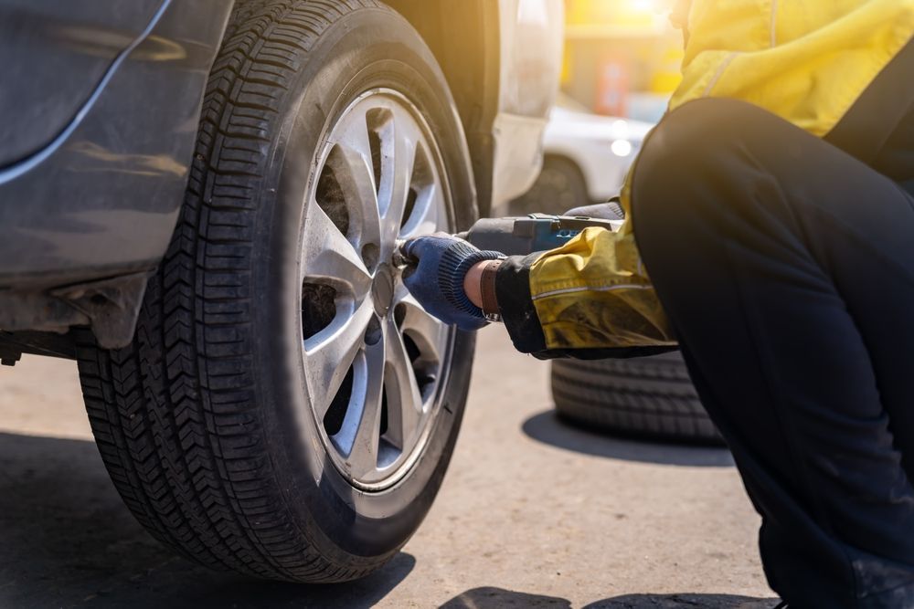 A worker in a yellow jacket and blue gloves uses an impact wrench to tighten a bolt on a car wheel.