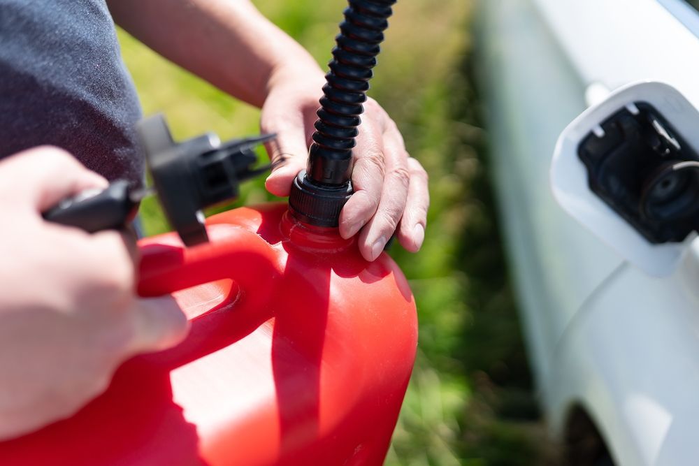 A person holds a red gas can with a black nozzle near the fuel filler door of a white vehicle.