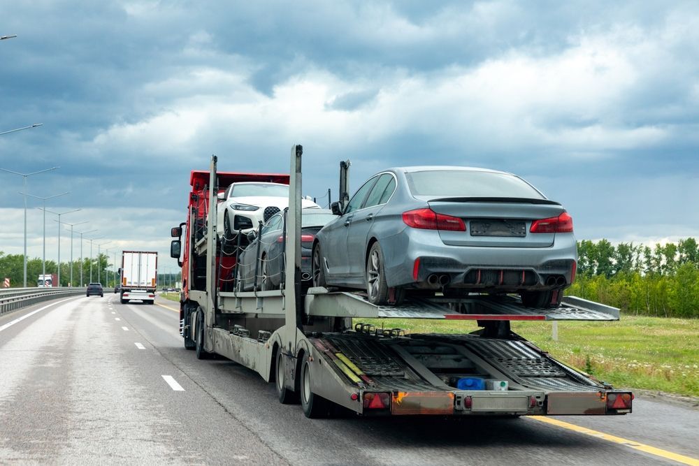 A car carrier truck transports several new luxury sedans along a multi-lane highway under a cloudy sky.