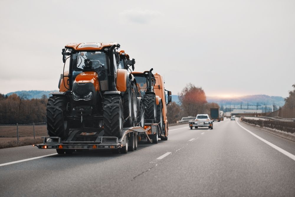 A flatbed truck transports two bright orange tractors along a highway during a sunset.
