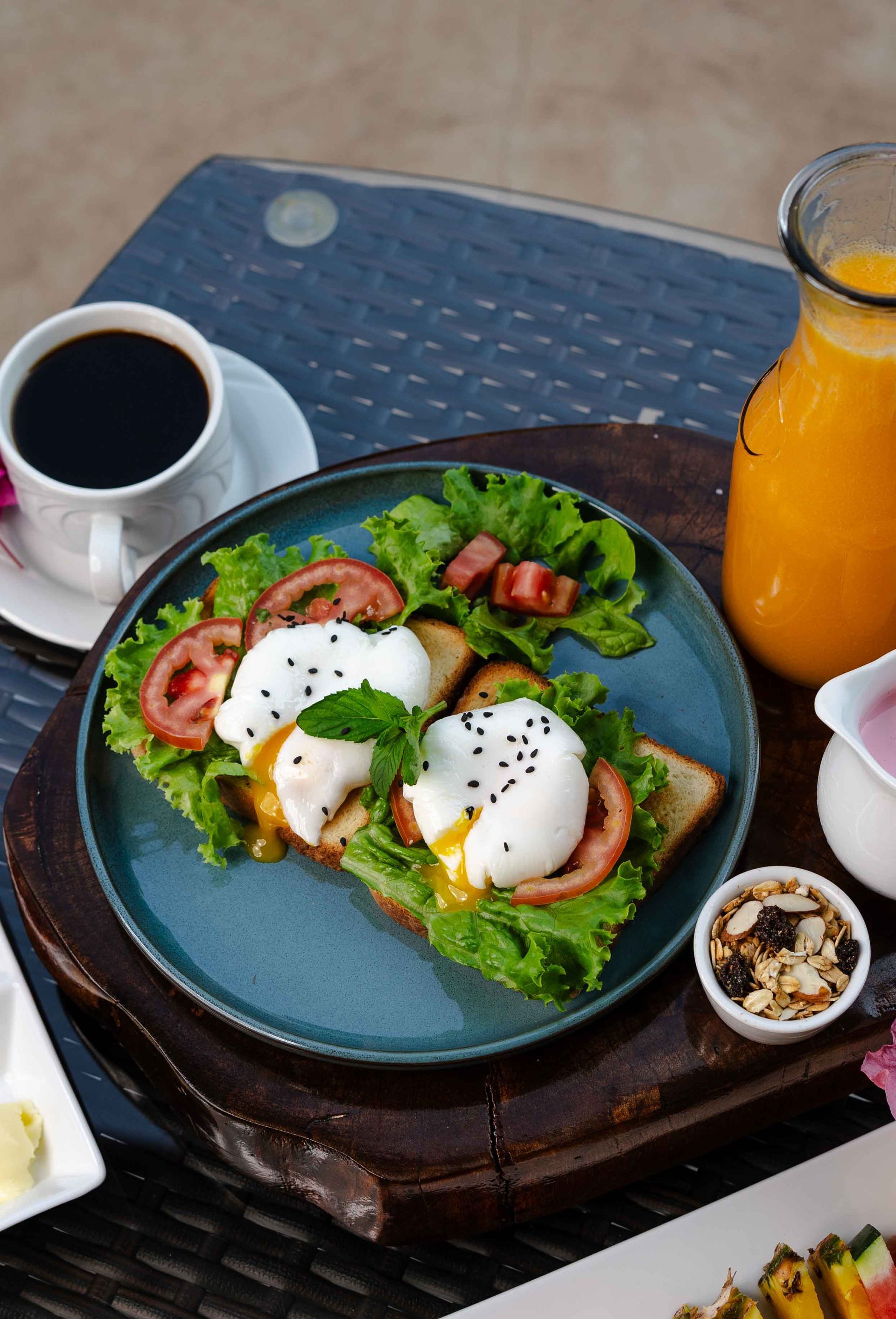 Desayuno: huevos escalfados sobre tostadas con café, jugo de naranja y granola en una bandeja de madera.