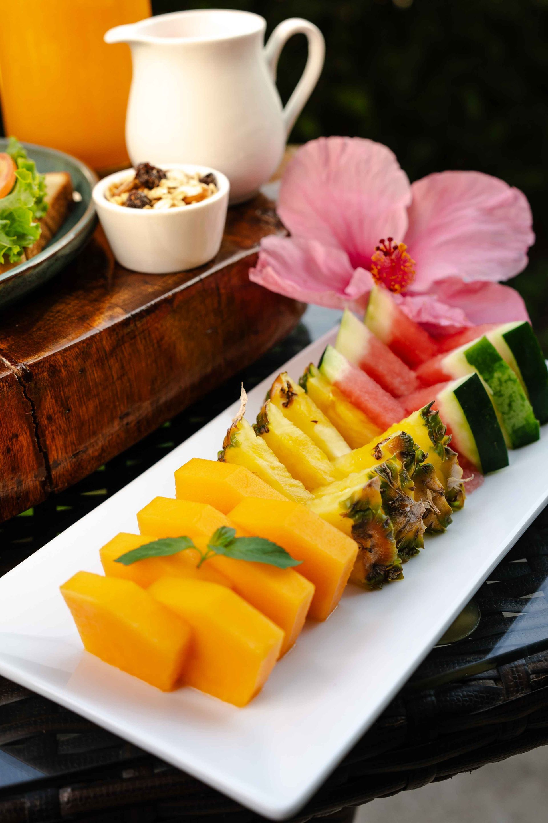 Plato de frutas con papaya, piña, sandía y pepino; una flor de hibisco y un pequeño plato de comida.