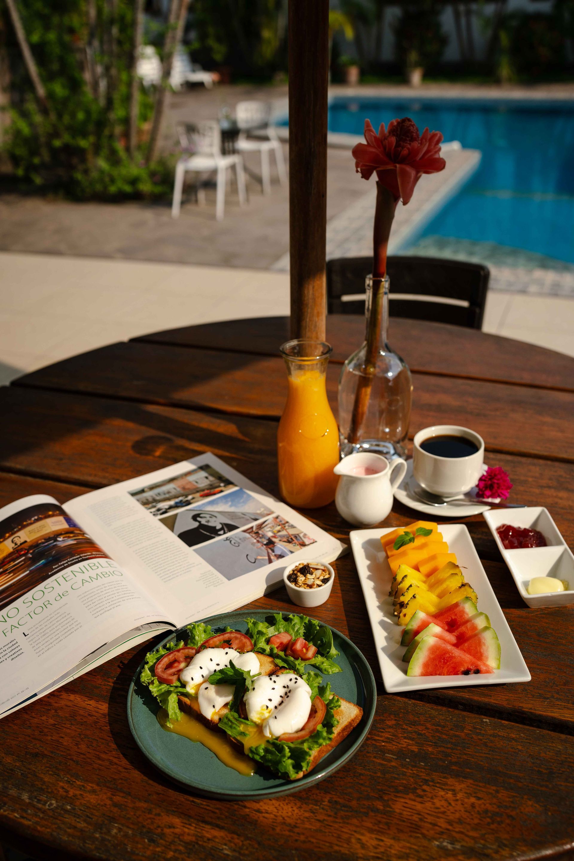 Mesa de desayuno junto a la piscina: tostadas, fruta, café, zumo y revista abierta.