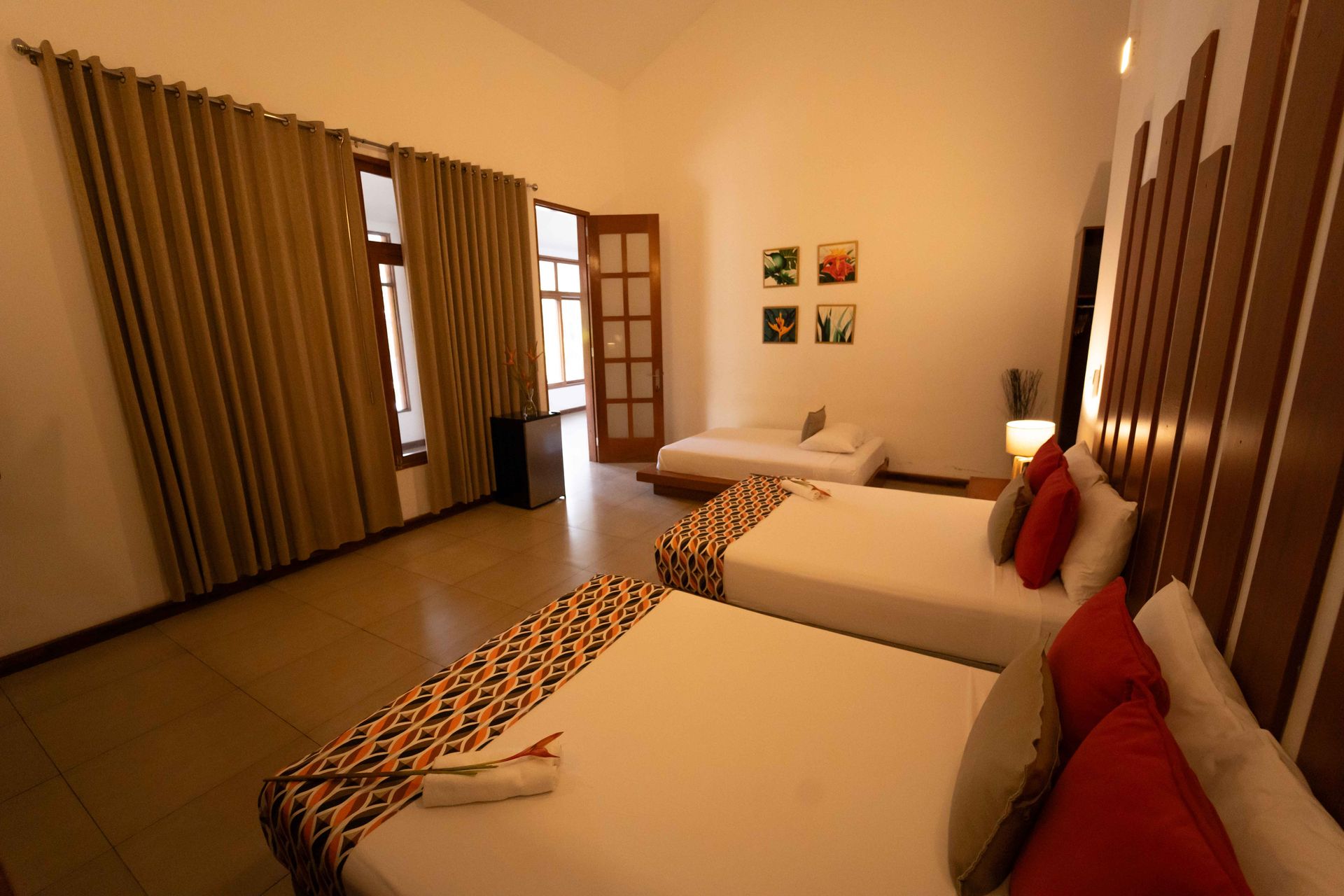 Dormitorio con tres camas, paredes neutras y una pared con acento de madera. La luz del sol entra a raudales por la ventana.