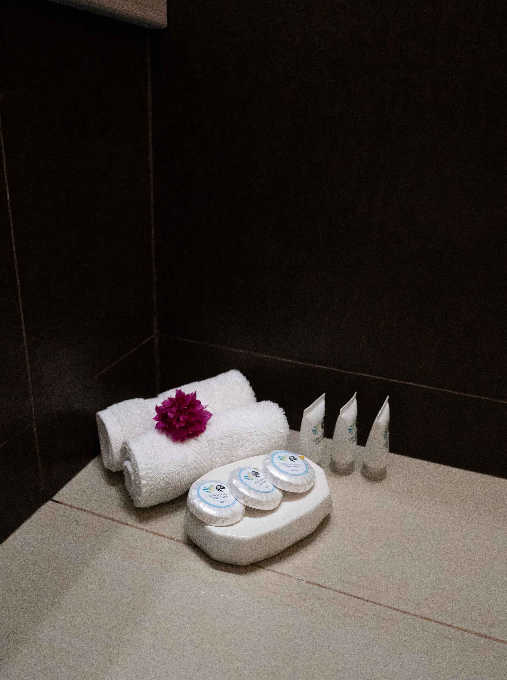 Toallas con flor rosa, jabones y artículos de tocador en un baño con azulejos oscuros.