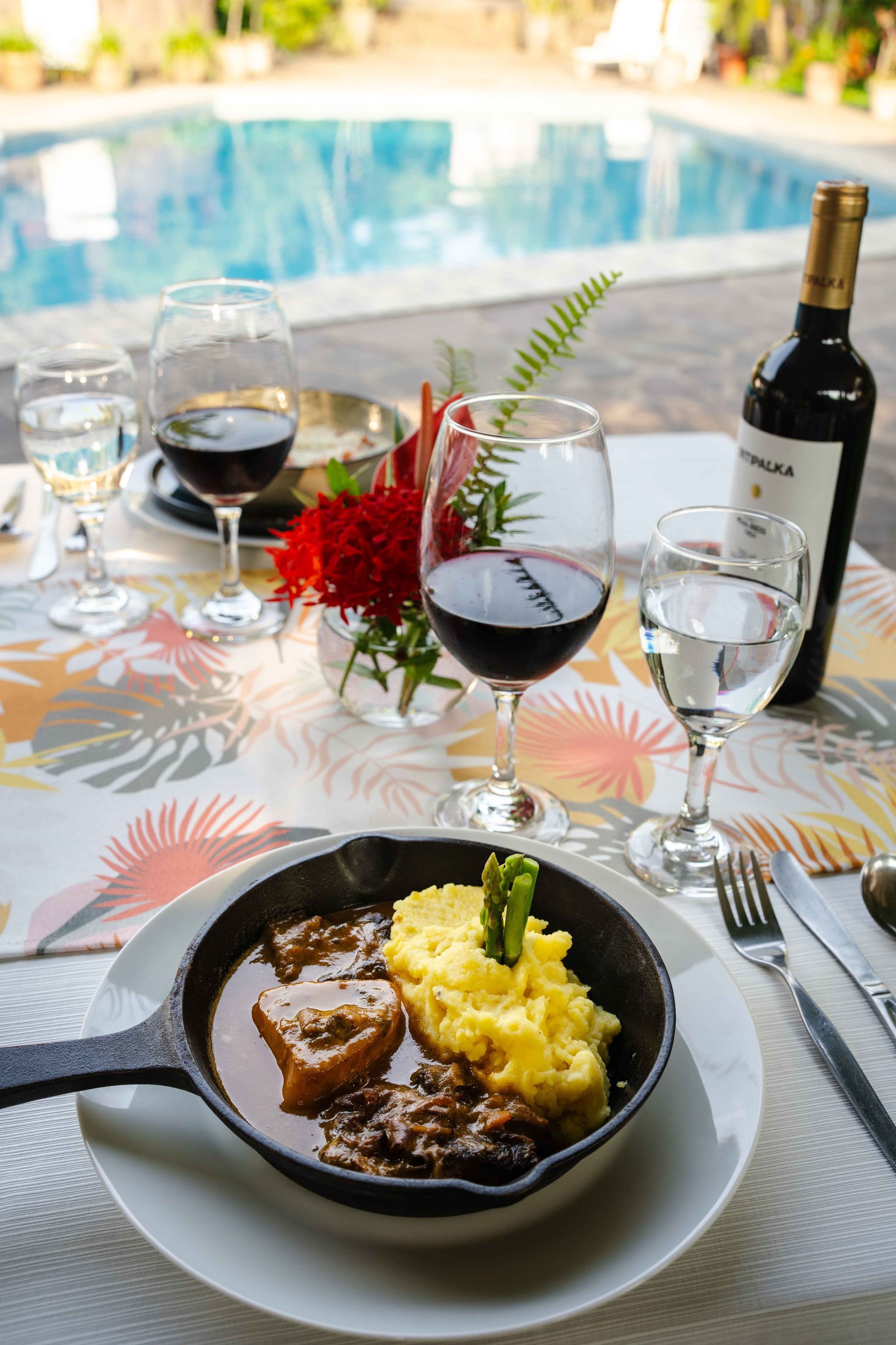 Una comida con vino tinto junto a la piscina: carne estofada y puré de patatas, con copas y una botella.