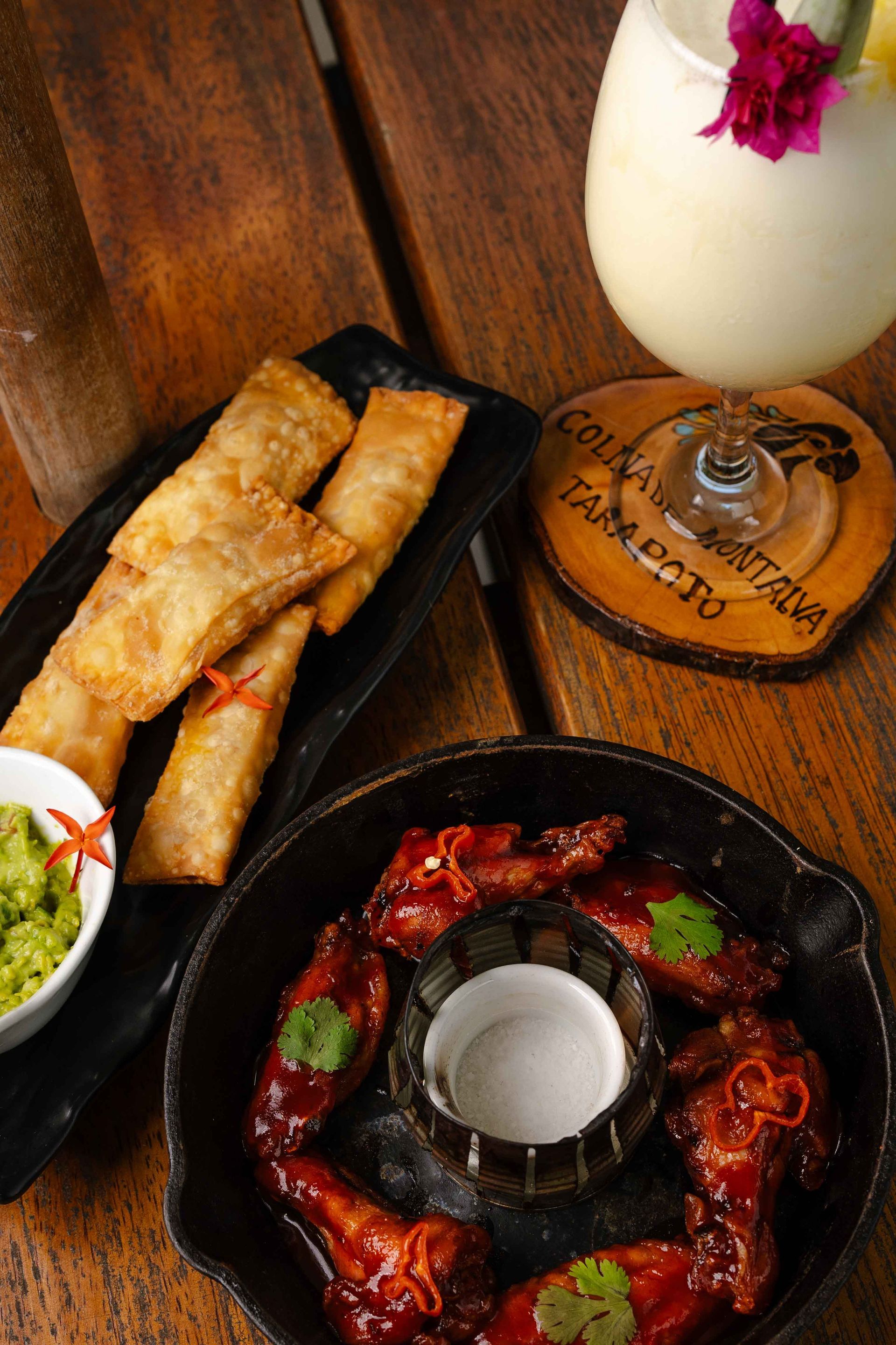 Comida frita y cócteles en una mesa de madera. Incluye alitas, rollitos de primavera, guacamole y una bebida.