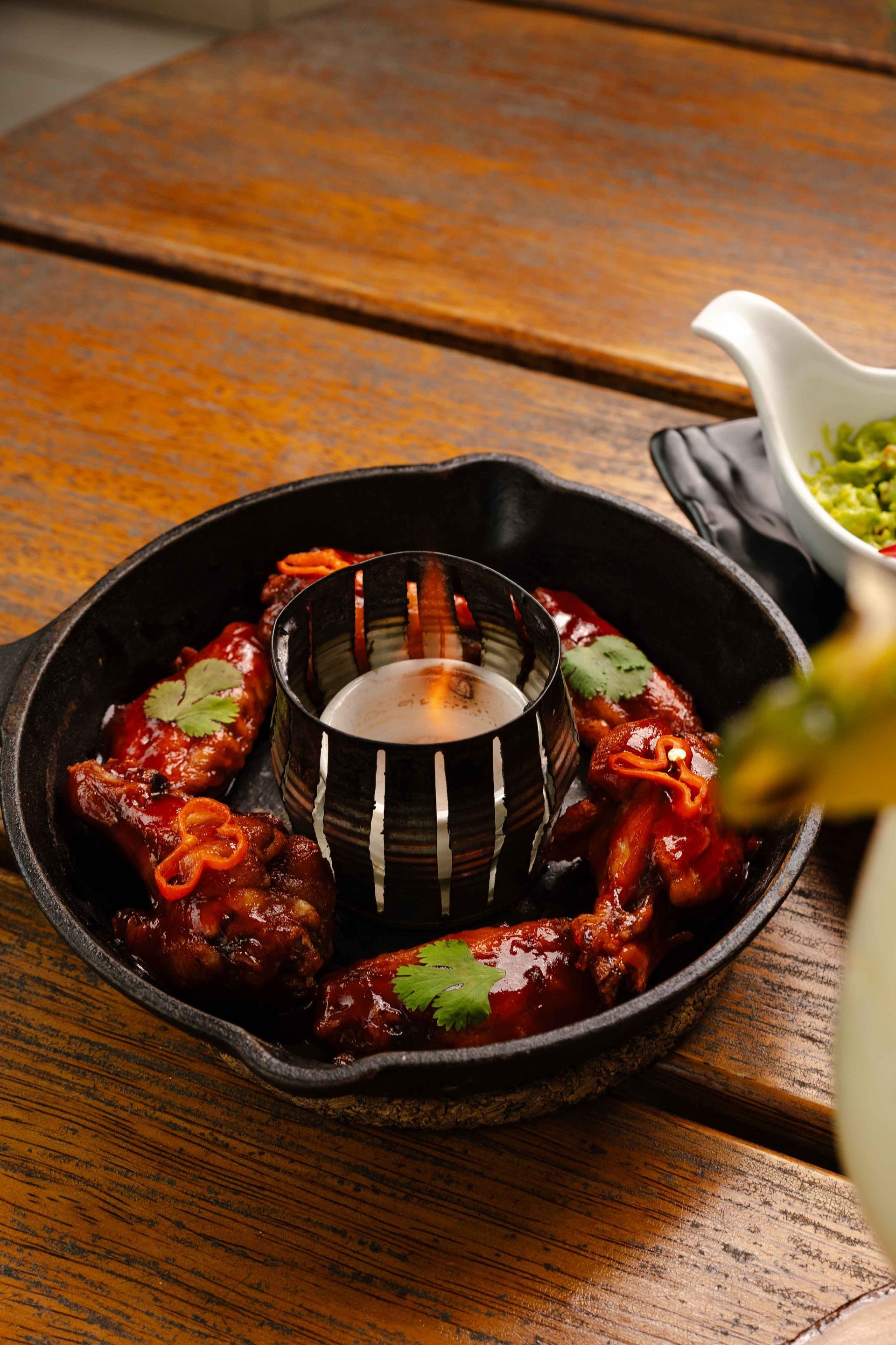 Alitas de pollo en una sartén de hierro fundido con un tazón pequeño de salsa y cilantro sobre una mesa de madera.