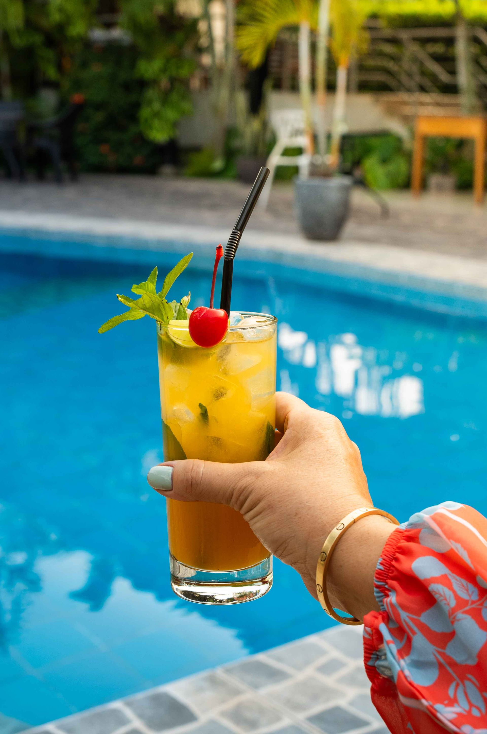 Mano sosteniendo un cóctel amarillo adornado con cereza y menta junto a una piscina.