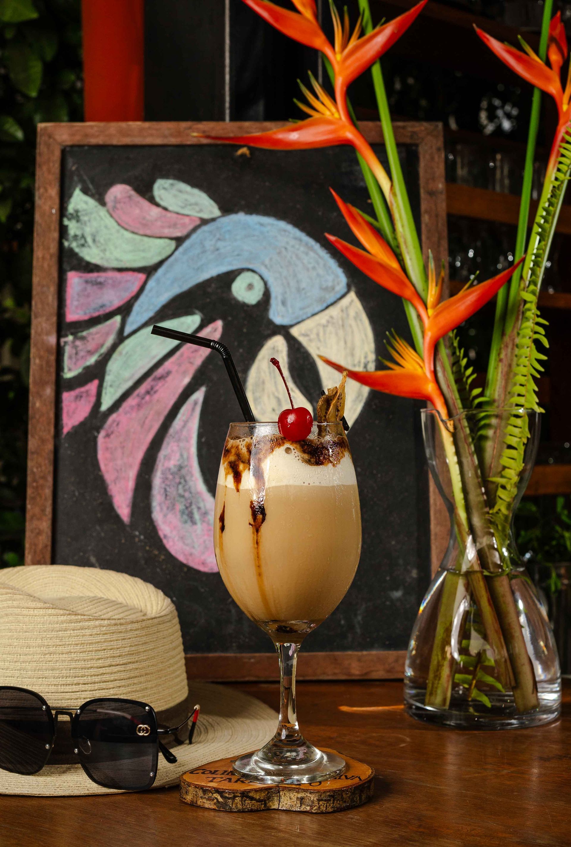 Cóctel con llovizna de cereza y chocolate, junto a un sombrero y gafas de sol, con un dibujo de un loro de fondo.
