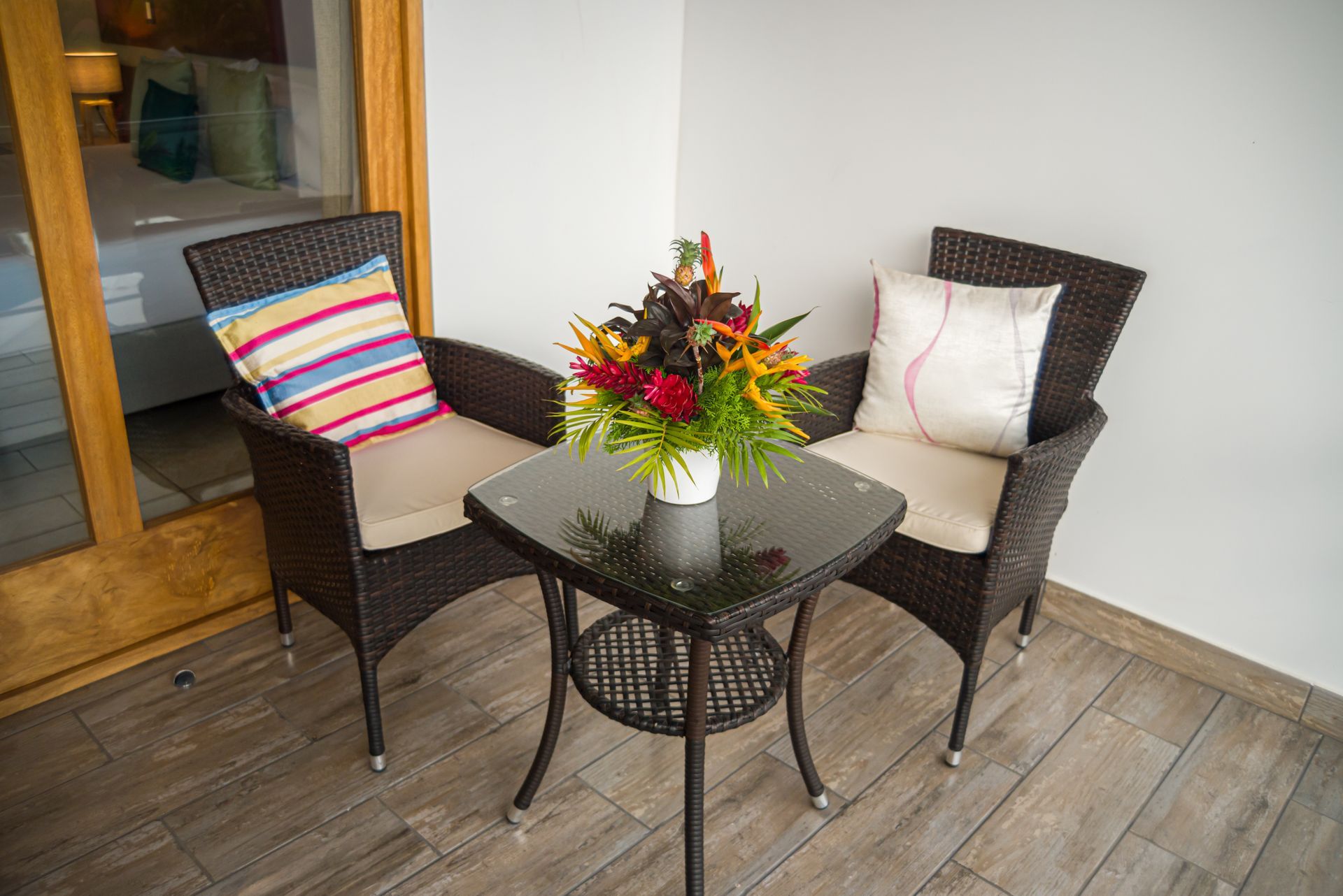 Dos sillas de mimbre con cojines y una mesa con flores en un balcón cerca de la puerta de un dormitorio.