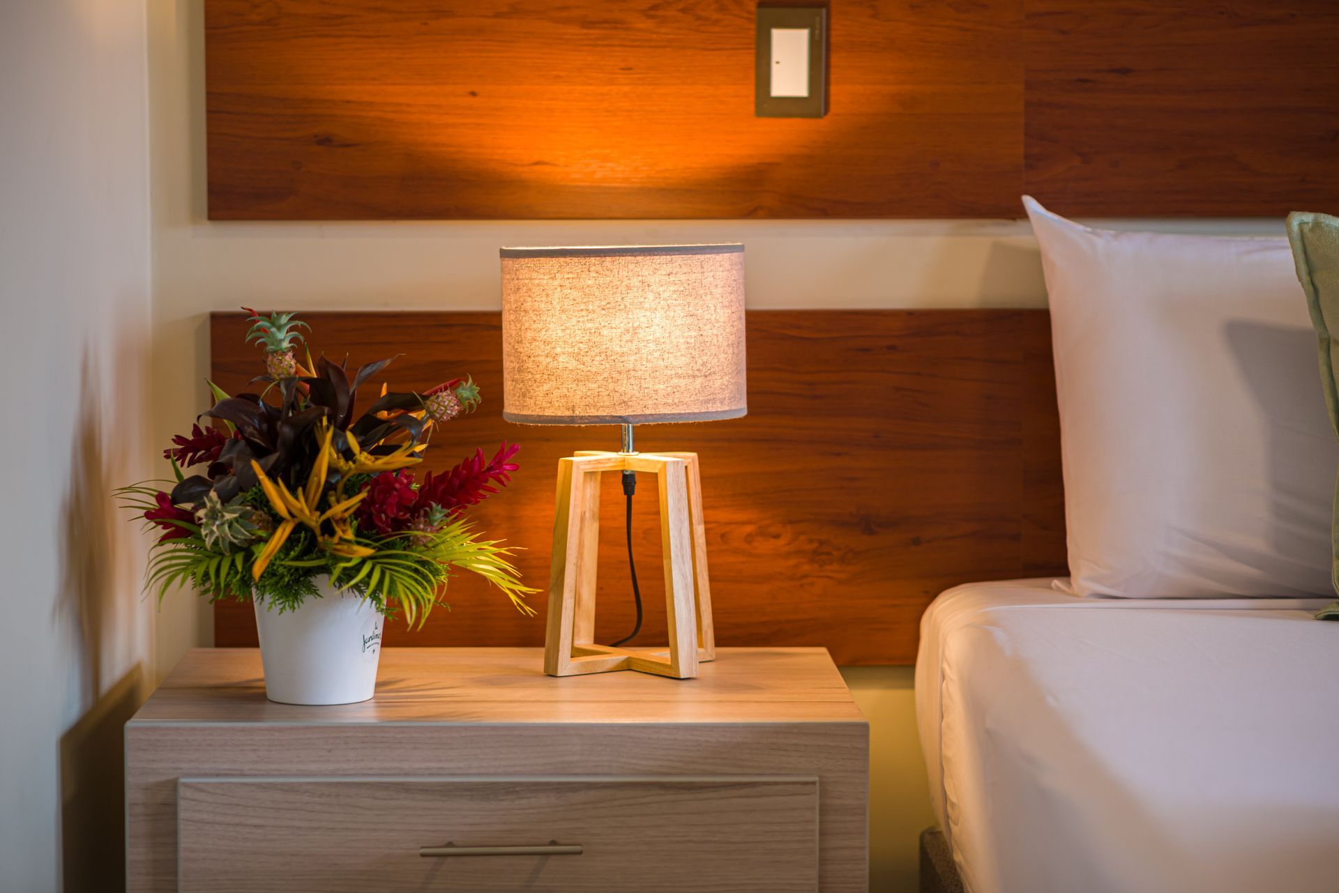 Mesita de noche con lámpara, arreglo floral y cama; luz cálida sobre cabecero de madera.