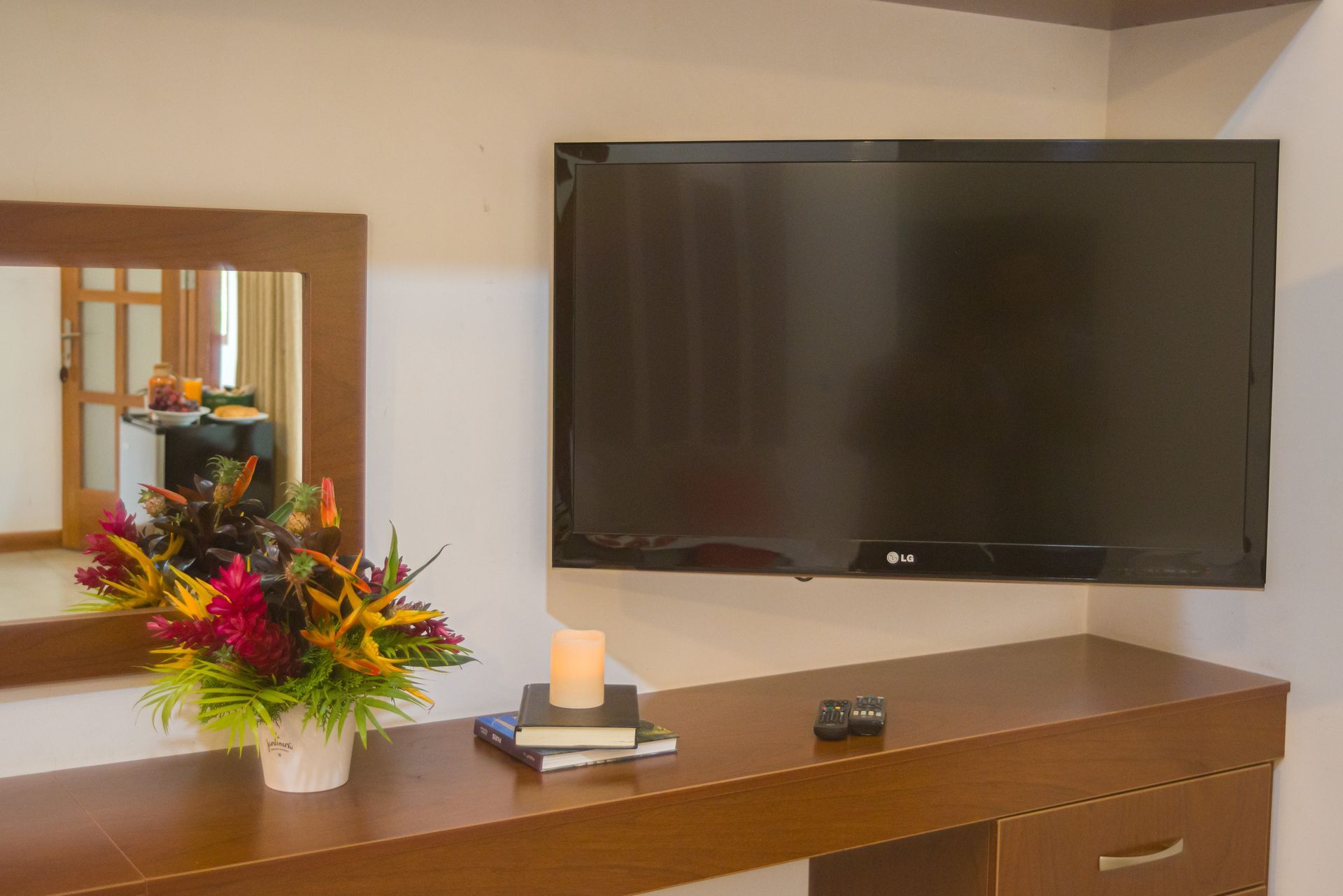 Un rincón de la habitación con un televisor montado en la pared sobre una consola de madera; un espejo, flores y libros.