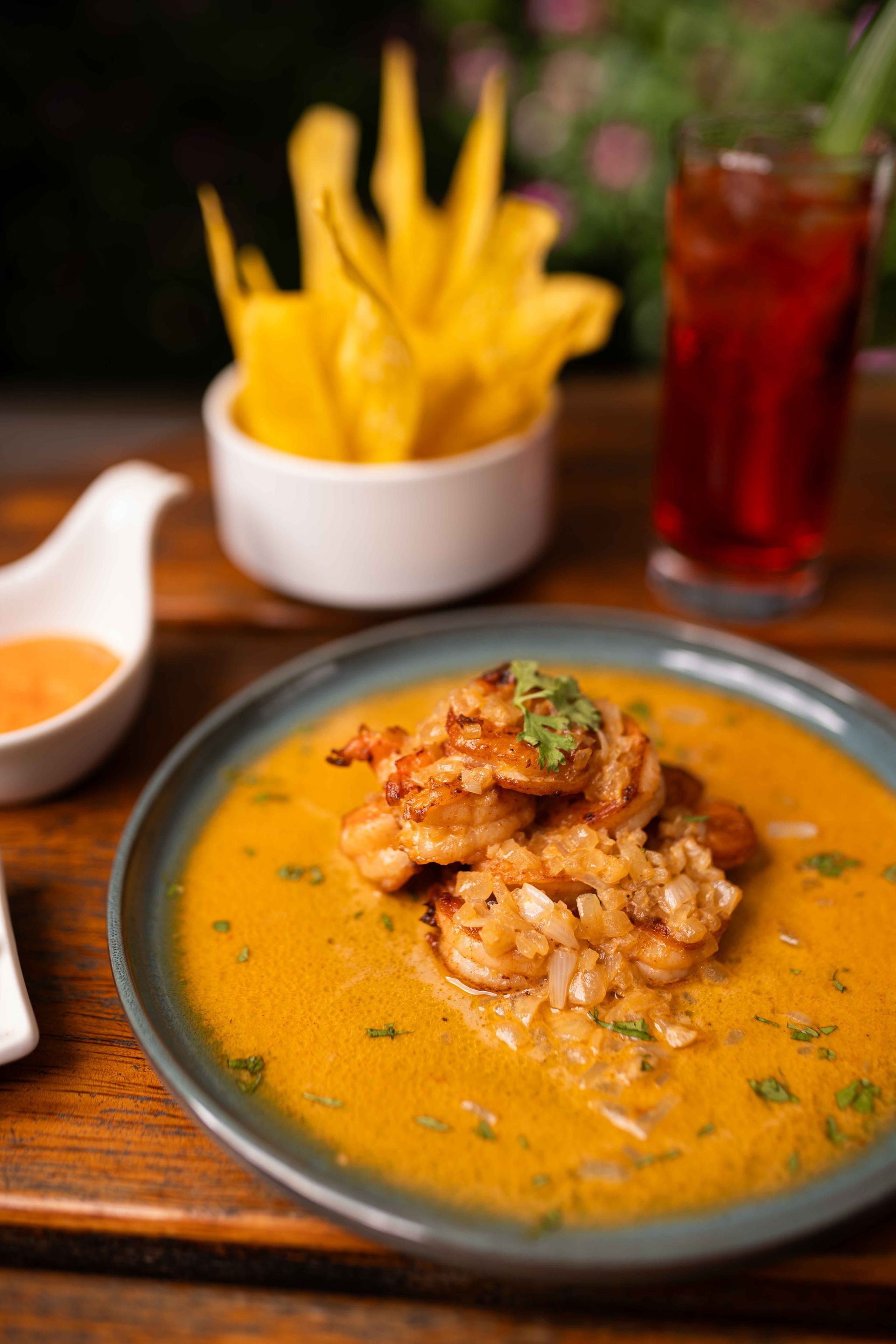 Camarones en salsa cremosa en un plato, con chips de plátano y una bebida, sobre una mesa de madera.