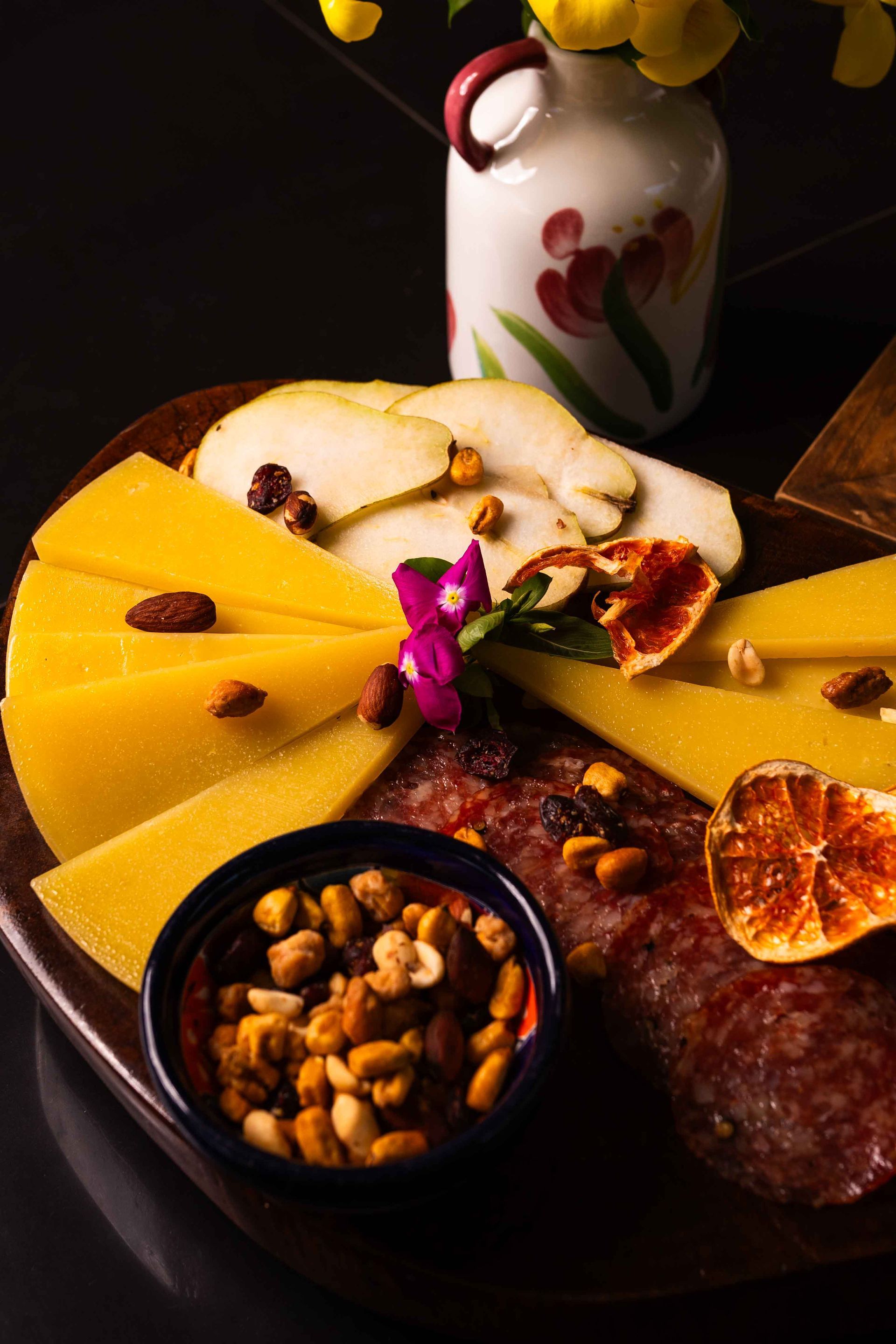 Tabla de embutidos con queso, salami, rodajas de pera, frutos secos y flores.