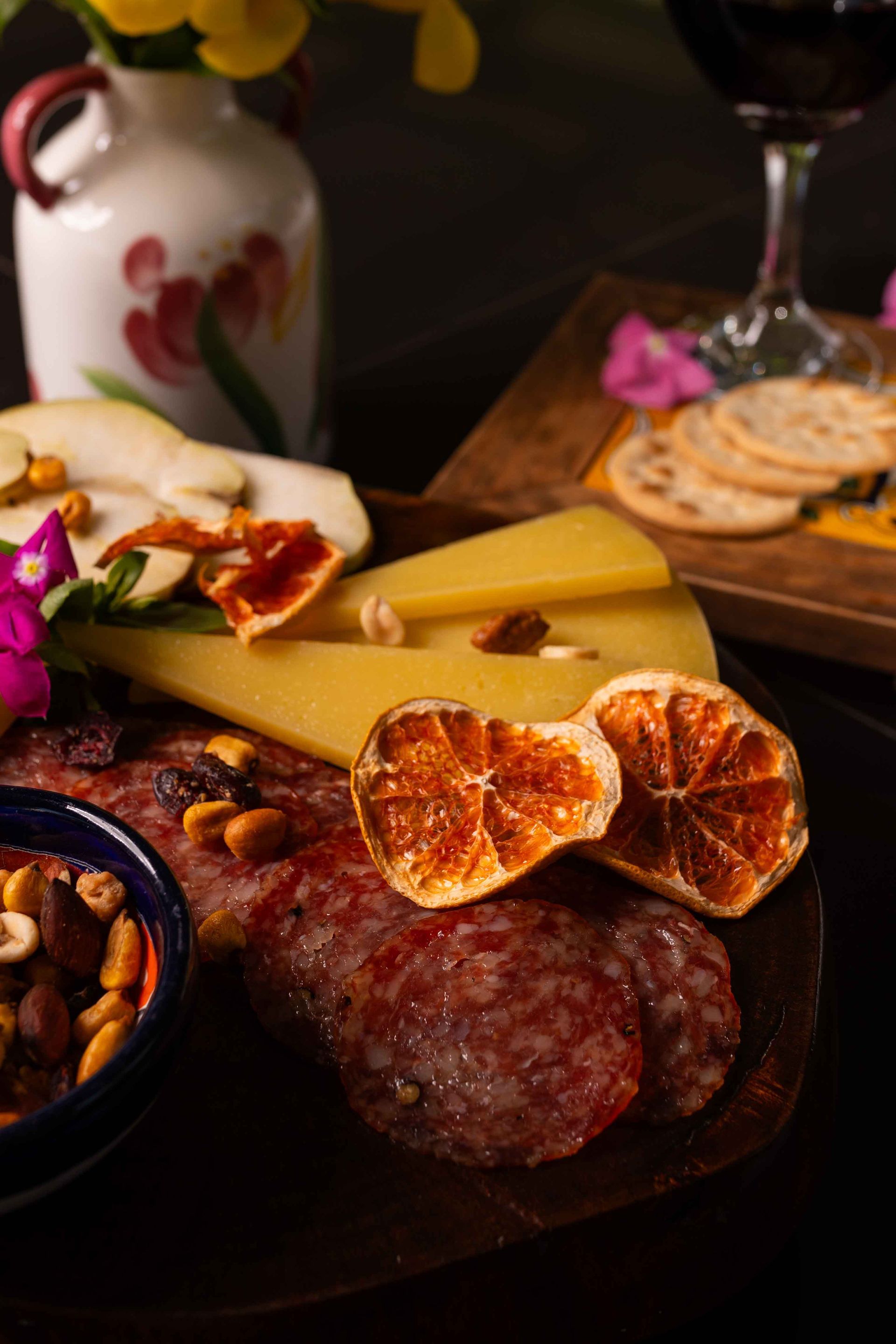 Tabla de embutidos con queso, carne, galletas y frutos secos, junto a una copa de vino y flores.