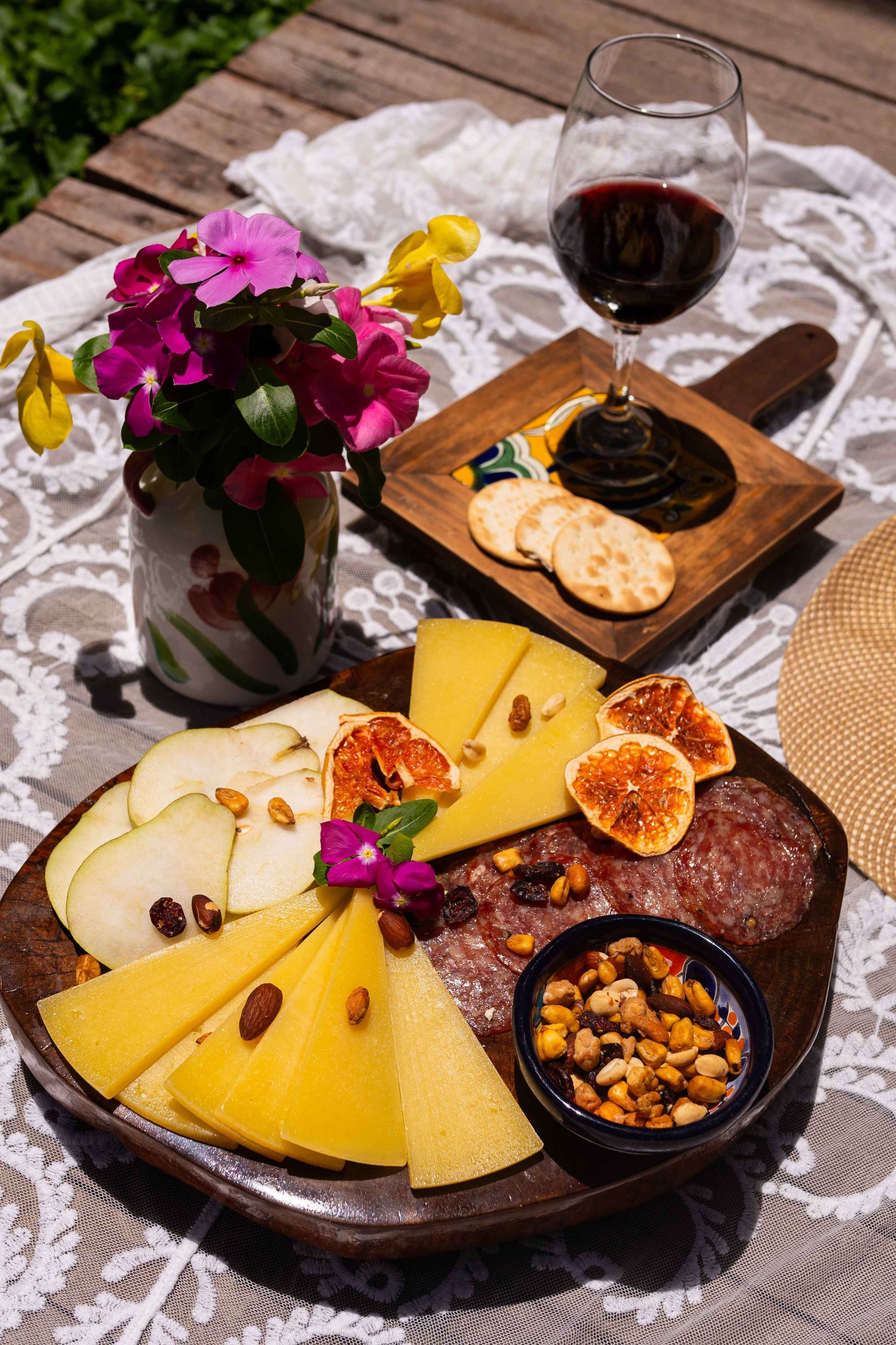 Tabla de embutidos con queso, fruta y frutos secos; copa de vino tinto, flores y sombrero de paja.