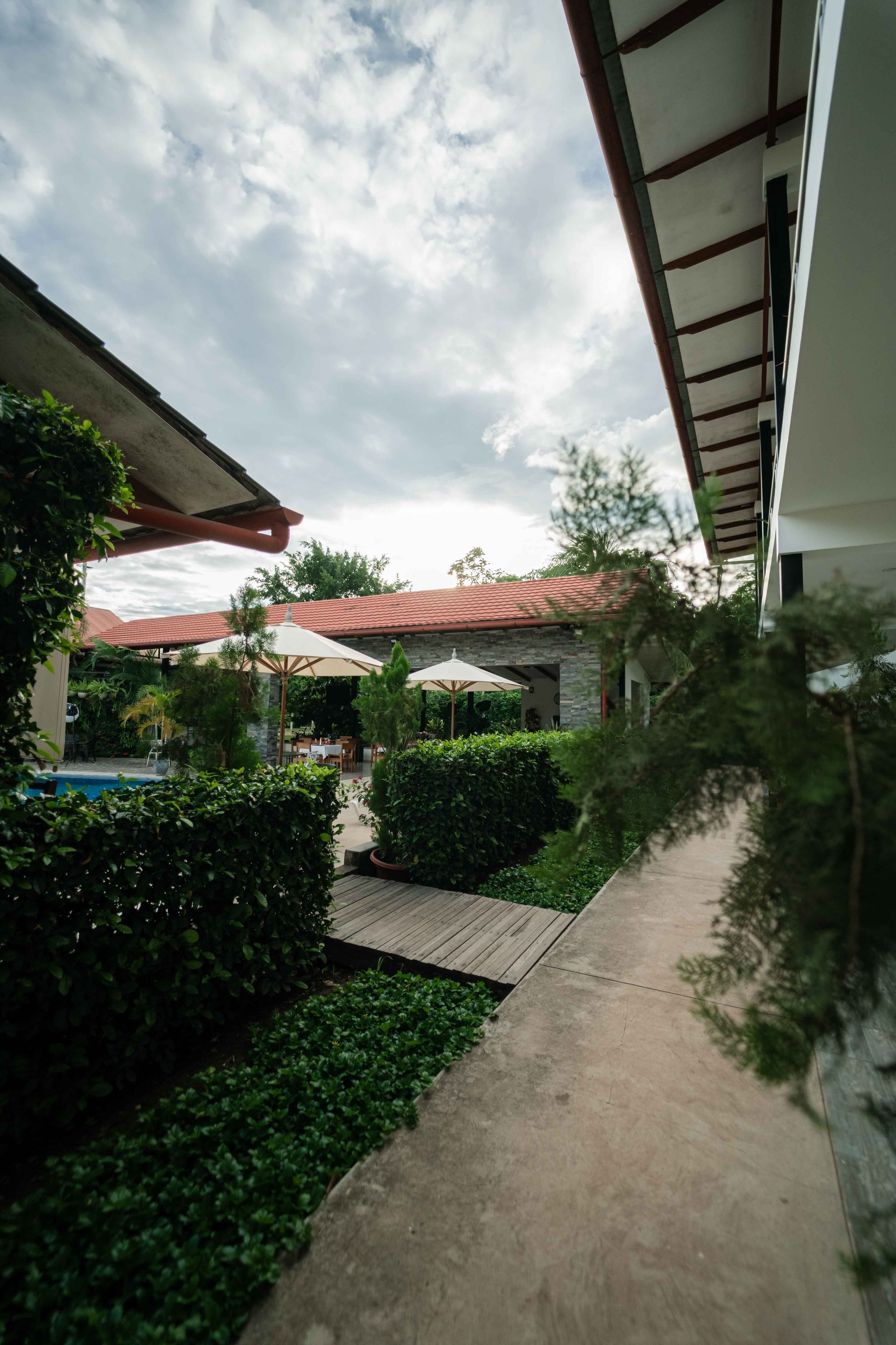 Camino entre edificios con vegetación, cielo nublado y destellos de zonas para sentarse.