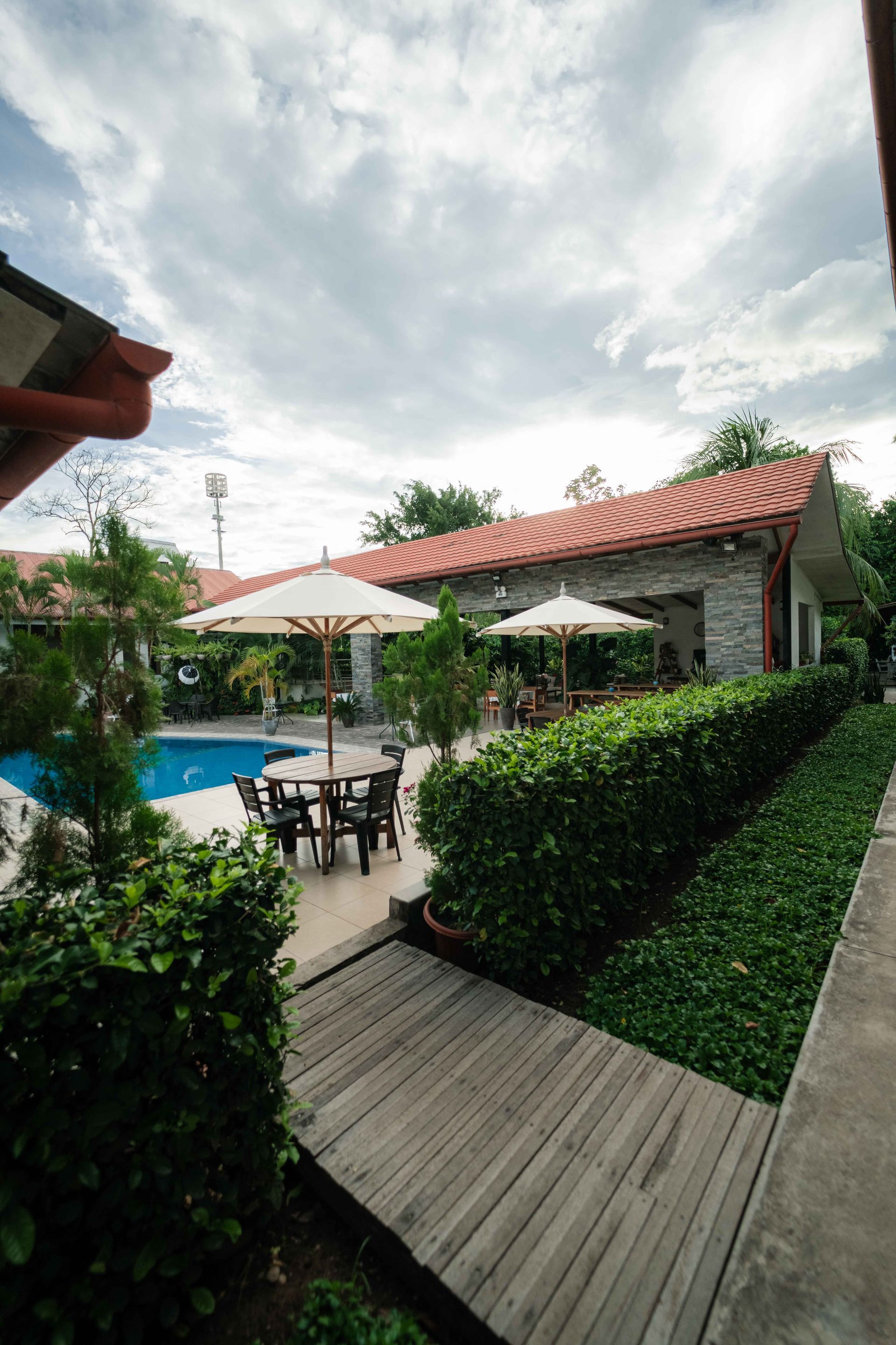 Patio exterior con piscina, mesas, sombrillas y setos verdes bajo un cielo nublado.