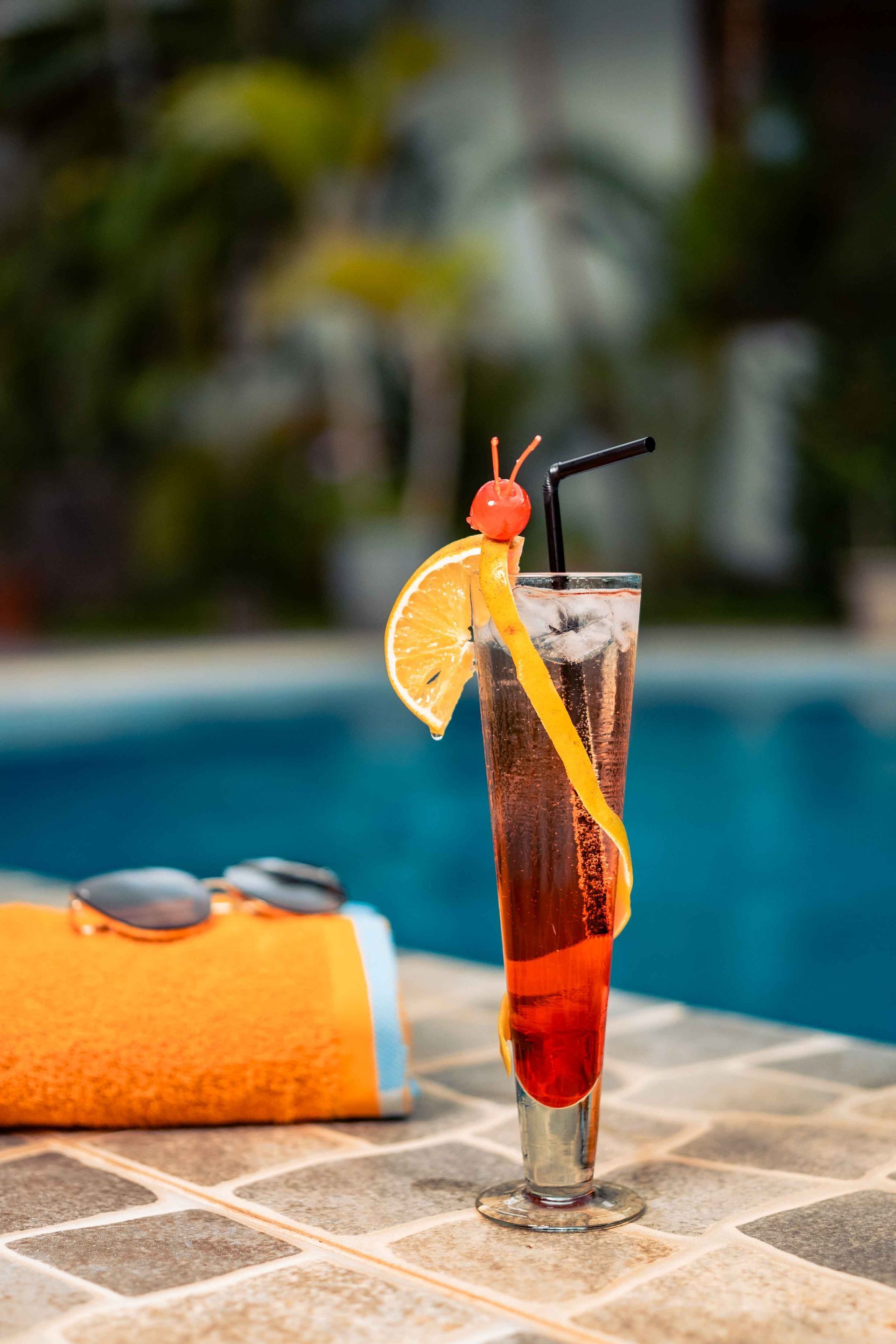 Cóctel con decoración de cerezas y limón junto a una piscina y toalla naranja con gafas de sol.