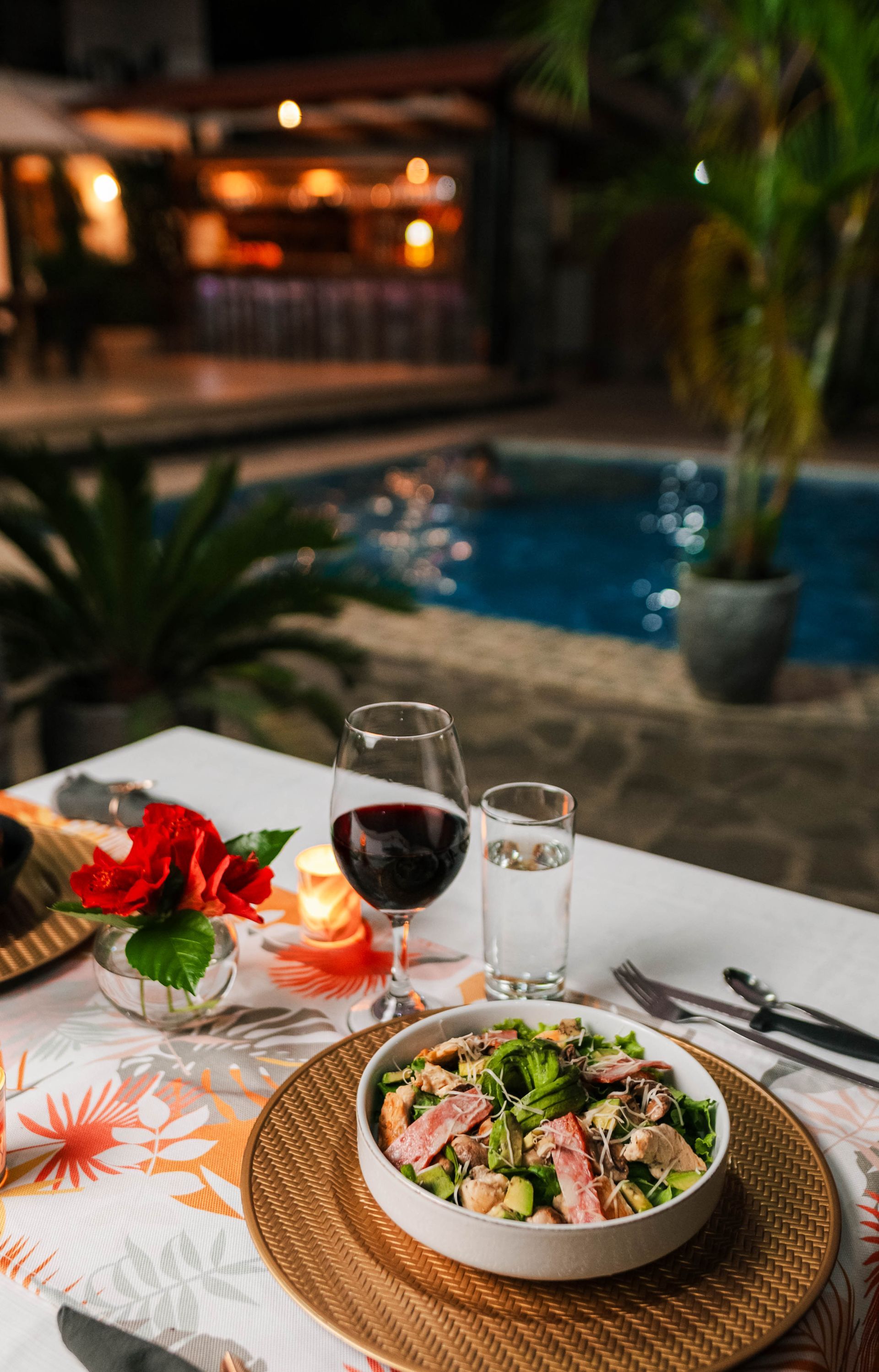 Cena romántica junto a la piscina: ensalada, vino tinto, agua, flores y una vela encendida sobre una mesa blanca.