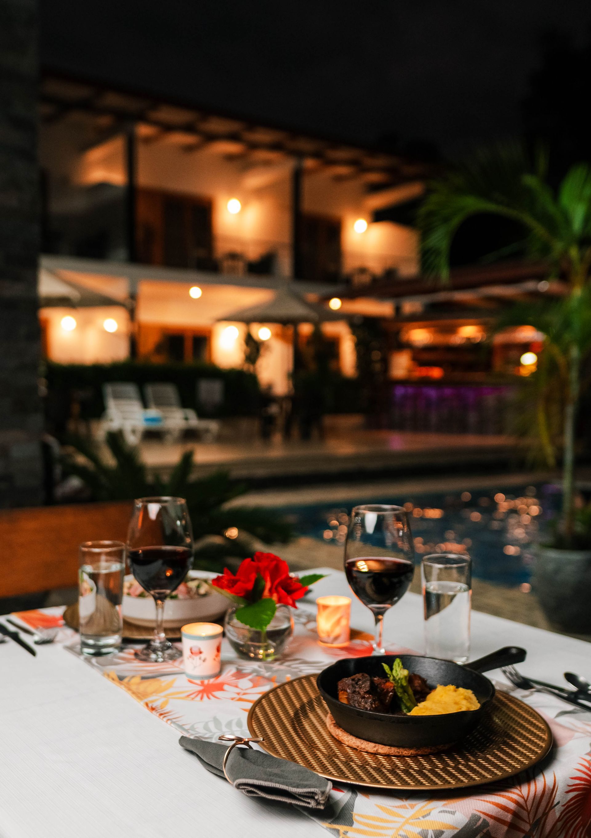 Cena romántica al aire libre: mesa puesta con vino, comida y flores; fondo borroso del edificio iluminado y la piscina.