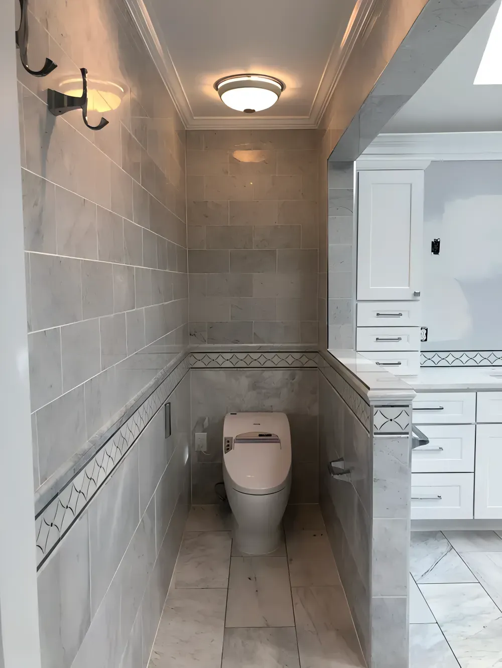 Narrow bathroom with gray tile, toilet, and overhead lighting, adjacent to a kitchen with white cabinets.