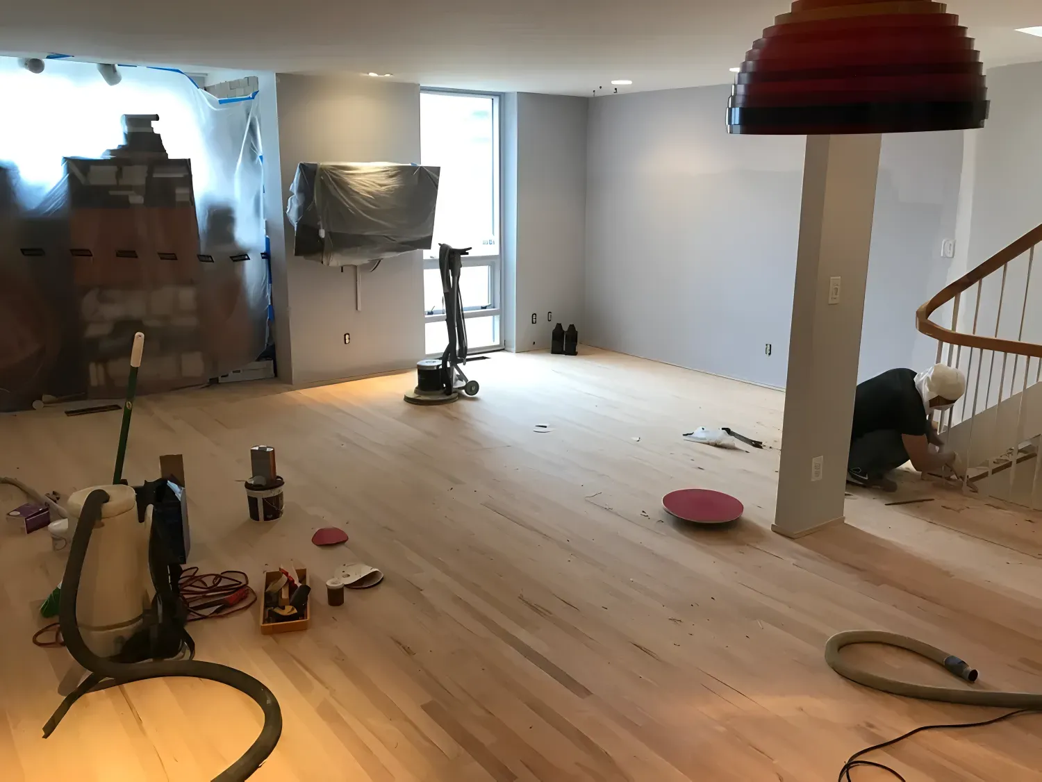 Interior view of a room under construction, with sanded wood floor, fireplace, and a worker near stairs.