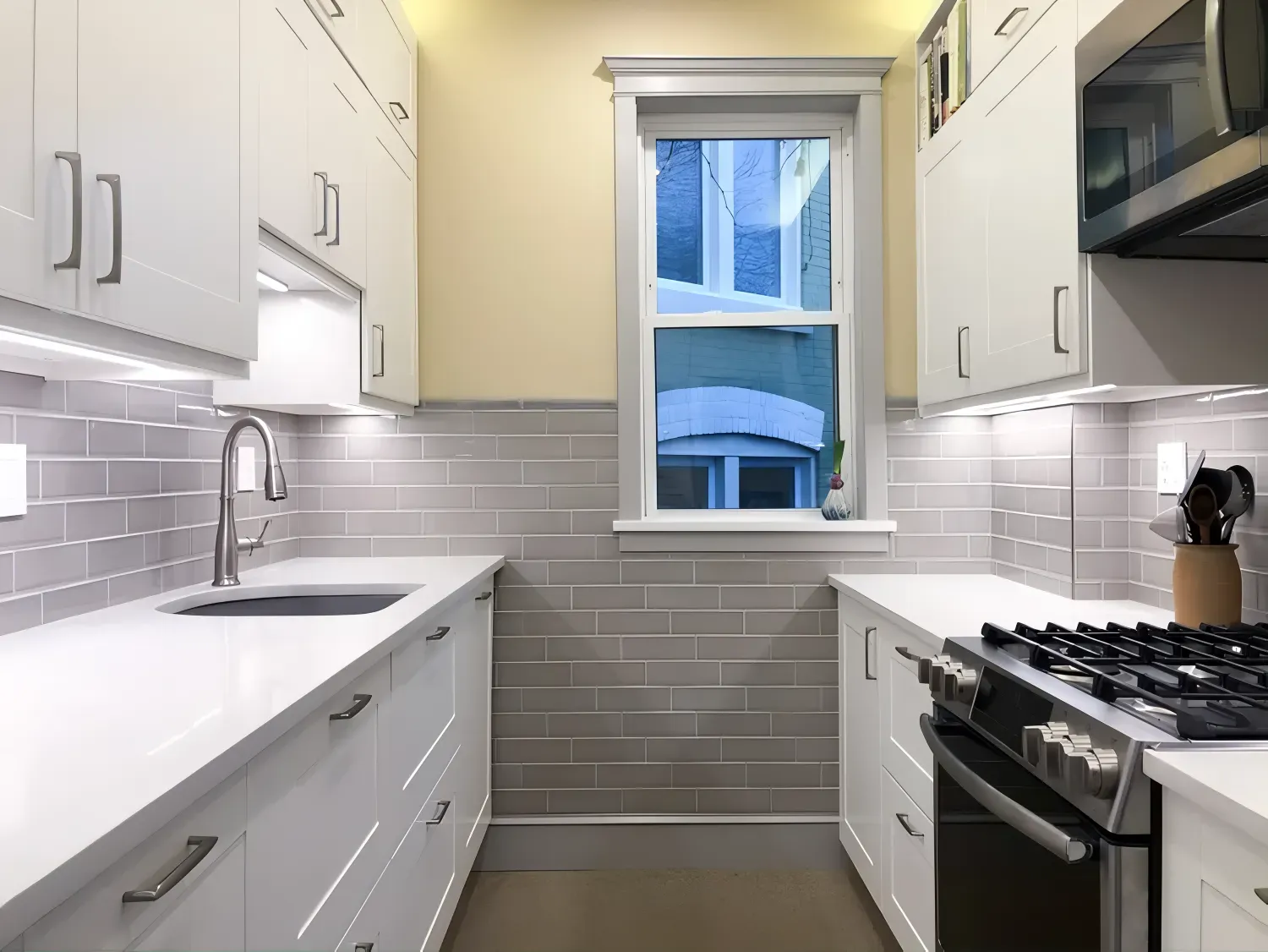 Narrow, modern kitchen with white cabinets, gray subway tile backsplash, and a window.