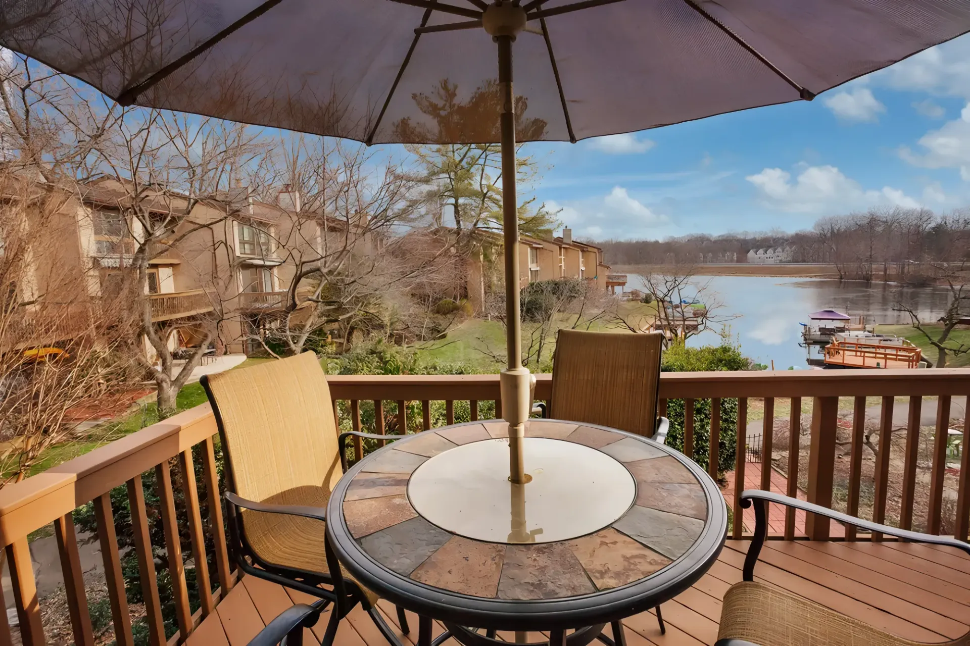 Deck with table, chairs, and umbrella overlooking a lake, townhouses in background, sunny day.