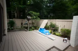 Wooden deck with construction materials against a beige fence and trees, daytime.