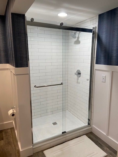 Shower with white subway tiles, glass door, and silver fixtures in a bathroom with blue wallpaper and white wainscoting.