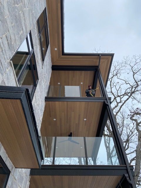 Modern multi-story building with a person on the balcony; grey stone facade, wood ceiling, and glass railings.