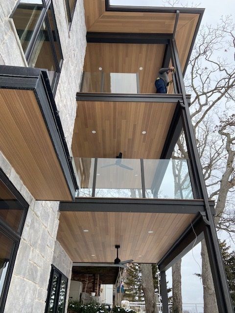 Exterior view of a modern house with wood-paneled balconies, glass railings, and a person working on the top balcony.