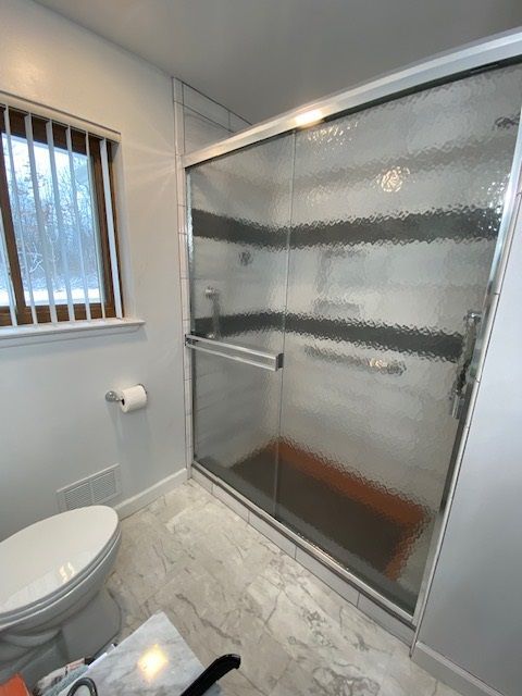 Bathroom with shower, toilet, window, and marble-like floor. The shower has a glass door and brown accent tiles.