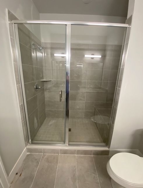 A shower with sliding glass doors, gray tiled walls and floor, and a toilet.